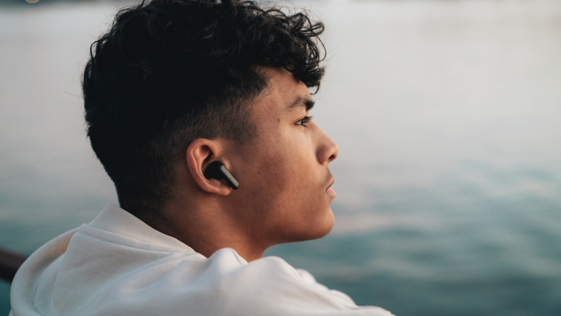 Widok z profilu młodego mężczyzny w białej bluzie z kapturem i czarnych słuchawkach, z wodą w tle.