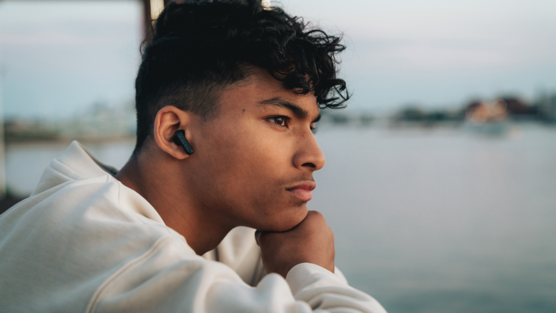 Młody mężczyzna w białym swetrze z ciemną słuchawką, zamyślony.