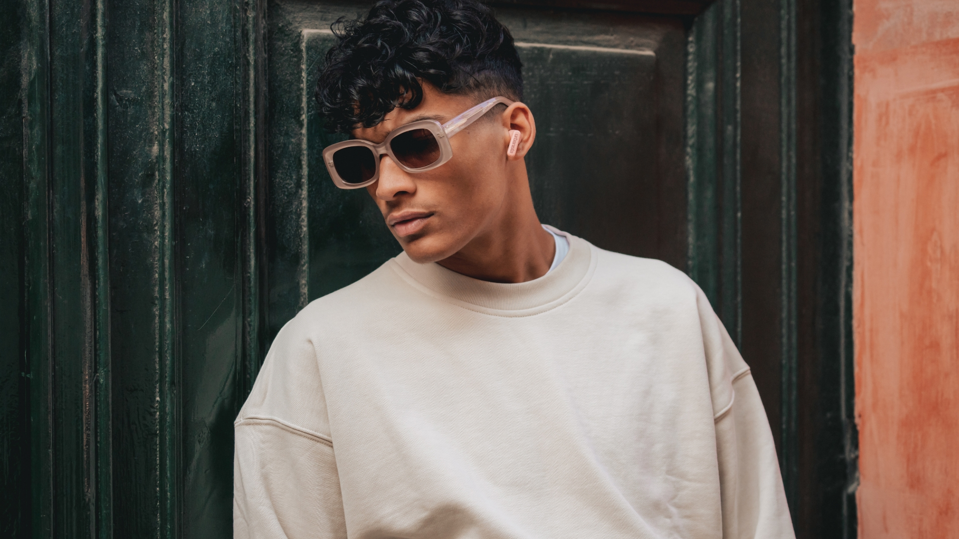 Mężczyzna w okularach przeciwsłonecznych i różowych słuchawkach opiera się o zielono-pomarańczową ścianę.