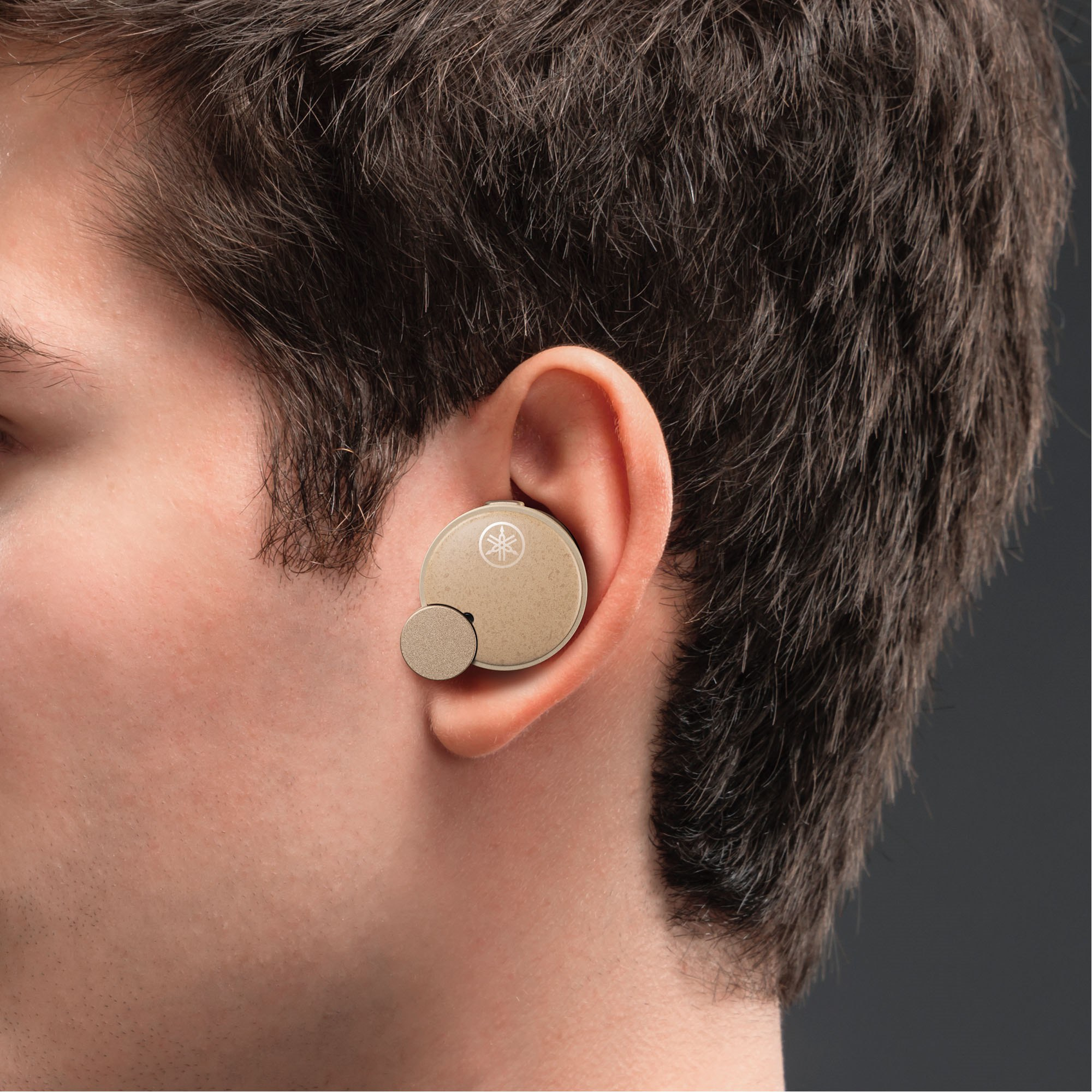 Ucho mężczyzny z włożonym beżowym bezprzewodowym wkładem dousznym, ciemne tło.