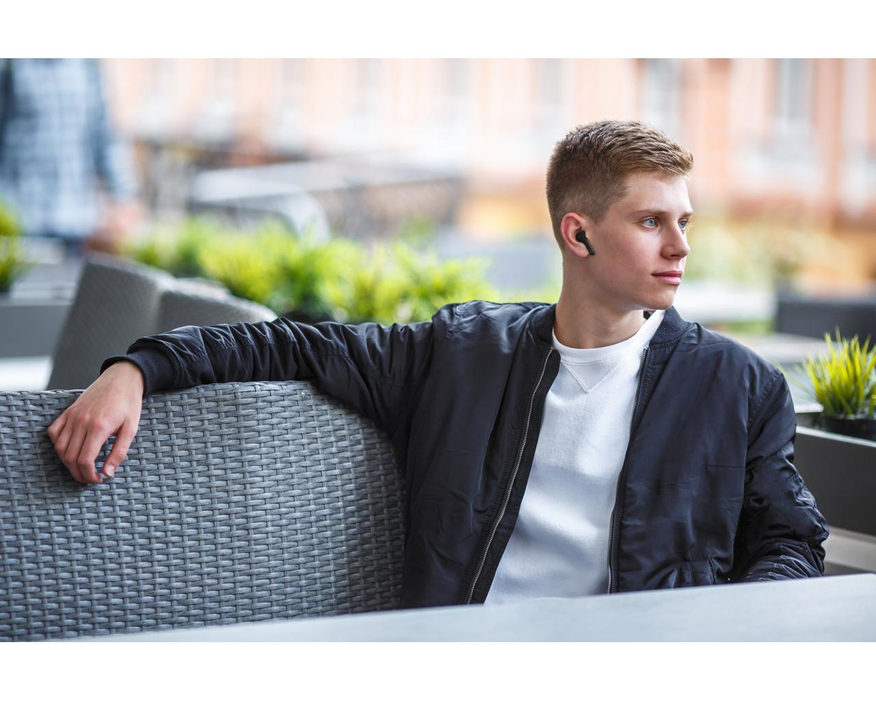 Młody mężczyzna w czarnej kurtce, słuchający muzyki z bezprzewodowymi słuchawkami, siedzący na zewnątrz.