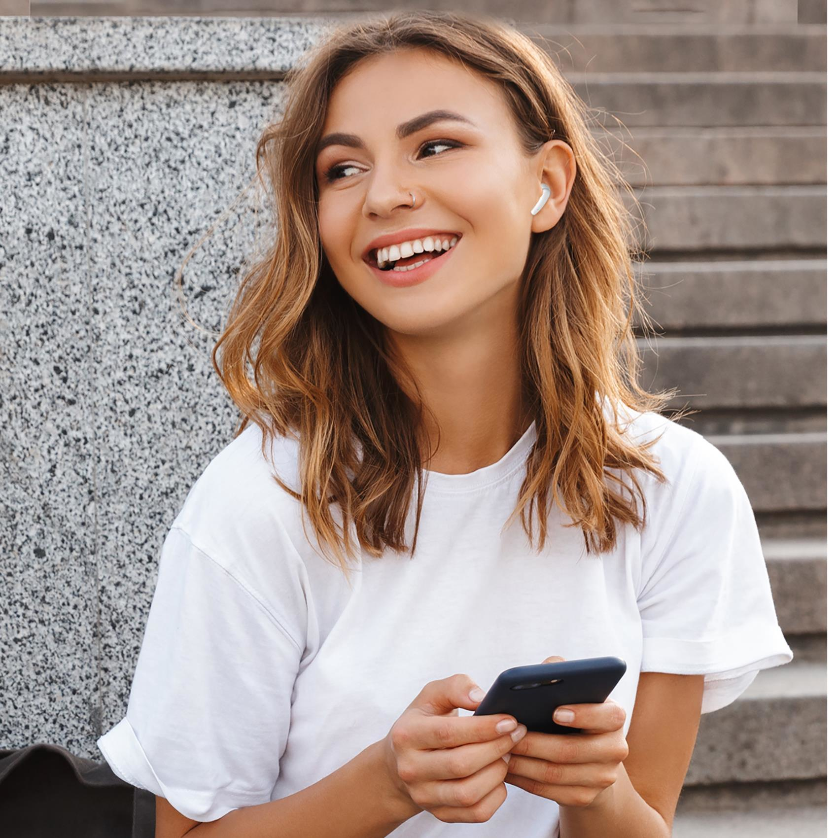 Uśmiechnięta kobieta, z słuchawką, trzymająca telefon.