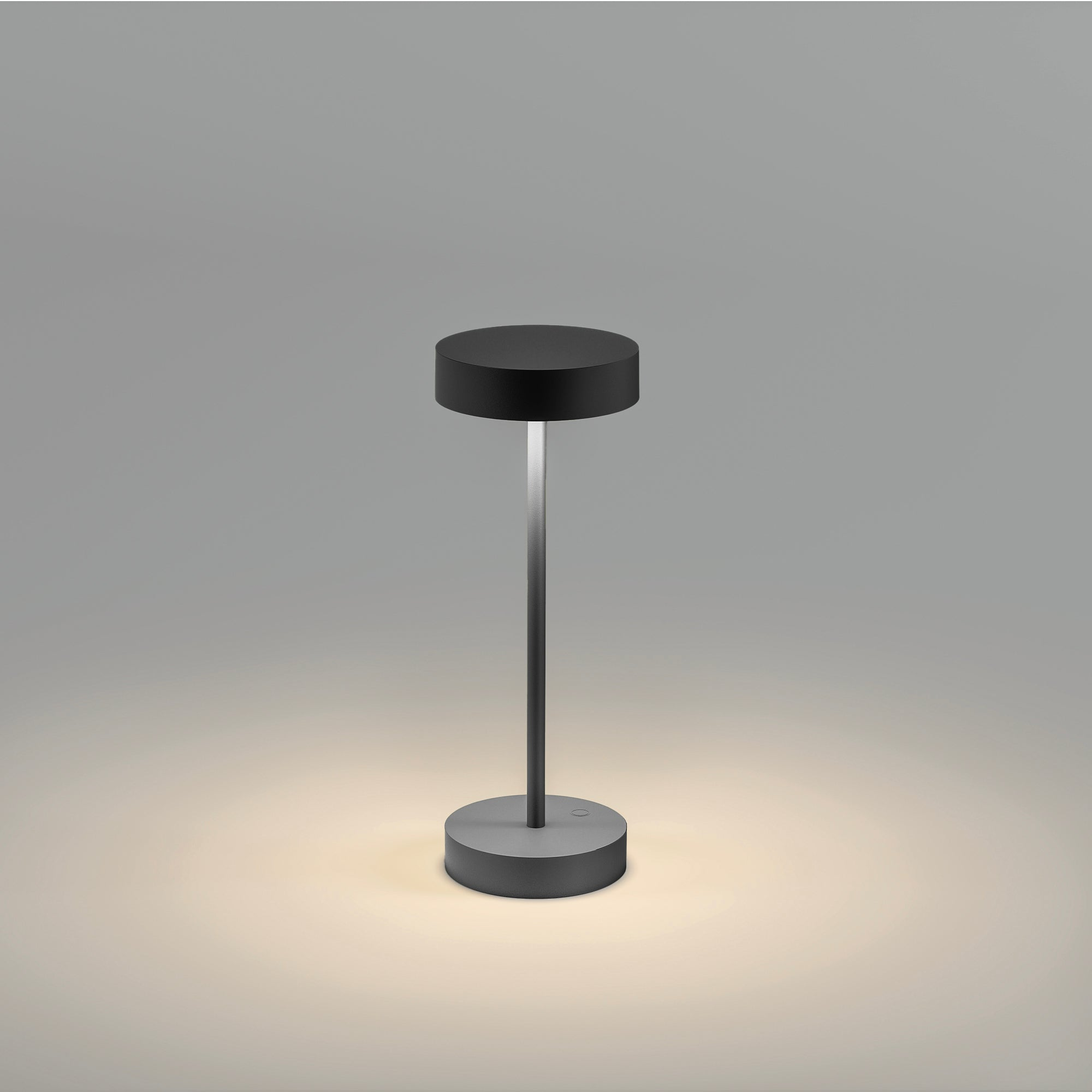 Eine schwarze Tischlampe mit kreisförmiger Basis und Oberseite, verbunden durch einen schwarzen Stiel. Licht ist an.