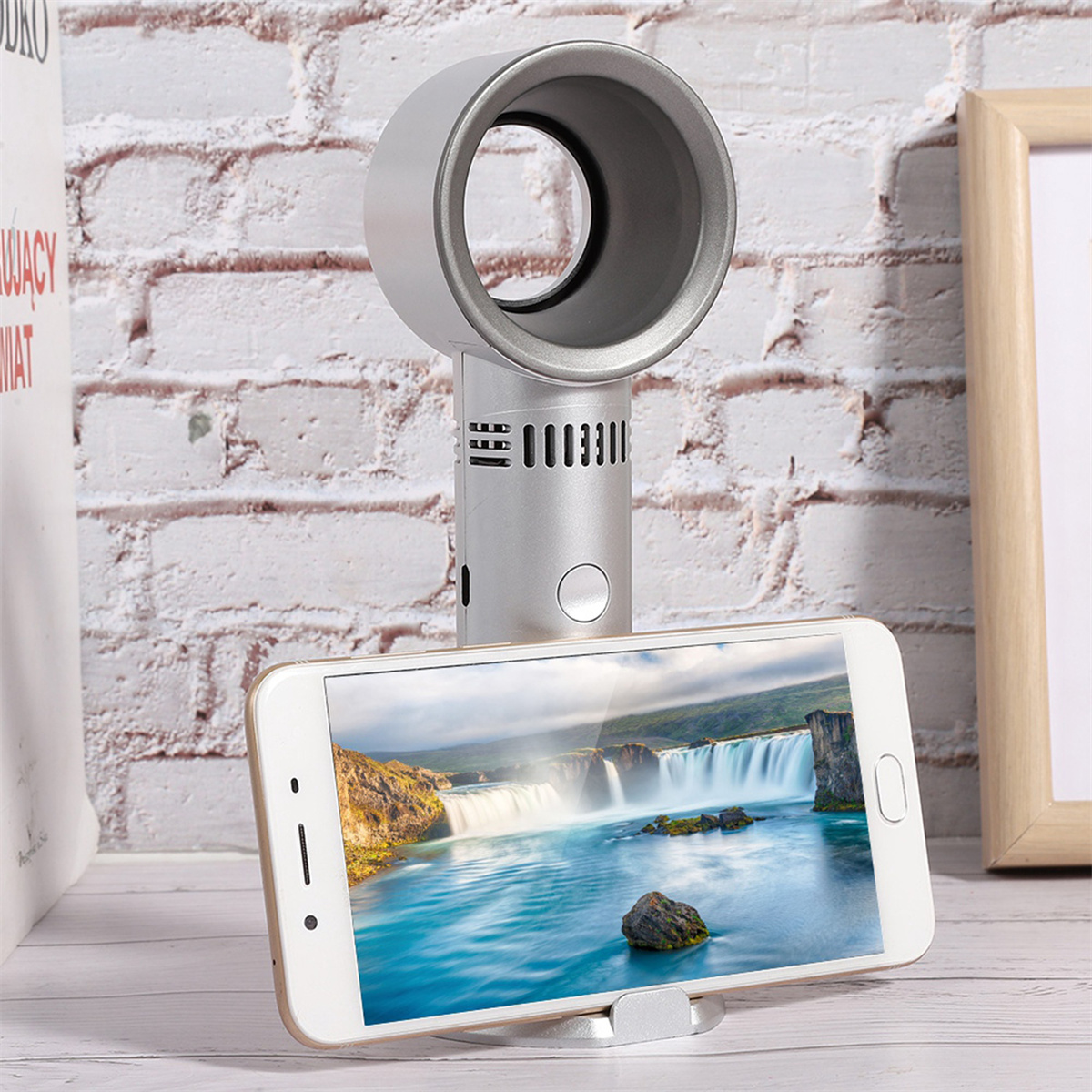 Silberner Ventilator mit Telefonständer, der ein Smartphone hält. Das Telefon zeigt eine Wasserfallszene. Ziegelwandhintergrund.
