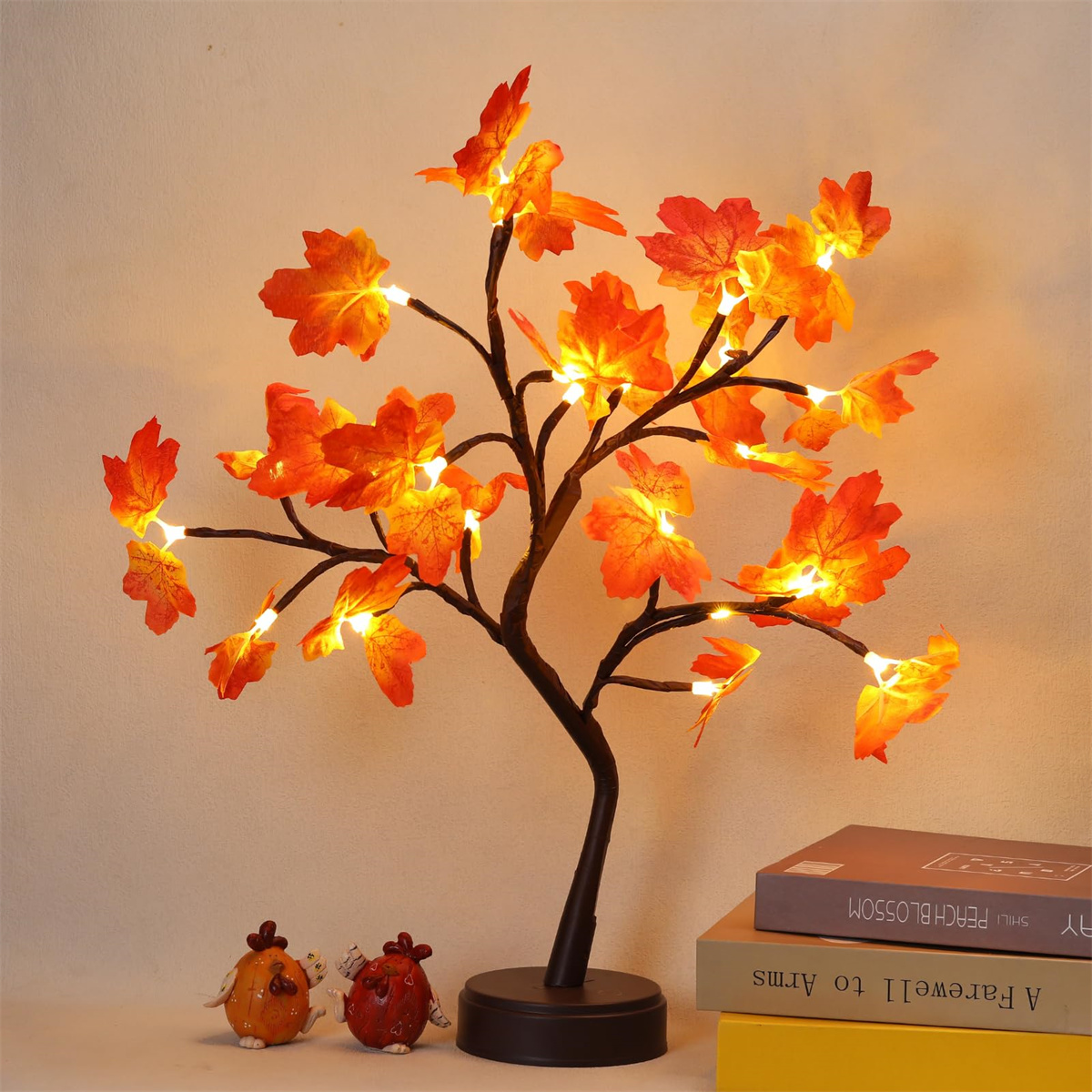 Künstlicher Baum mit orangefarbenen Blättern und Lichtern. Zwei kleine Hühnerfiguren und Bücher befinden sich im Vordergrund.