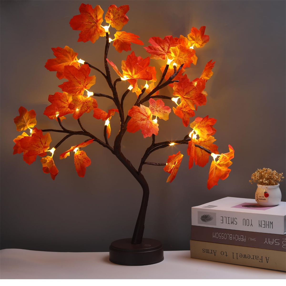 Ein dekorativer Baum mit orangefarbenen Blättern und kleinen Lichtern. Bücher und ein kleiner Topf im Hintergrund.