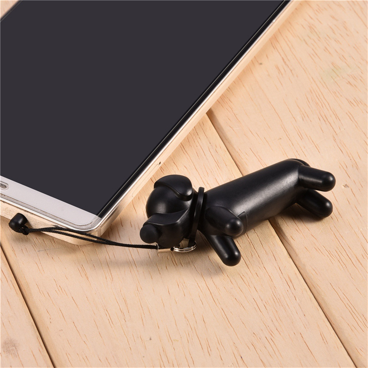 Schwarzes hundeformiges Telefonzubehör neben einem Smartphone. Es liegt auf einer Holzoberfläche.