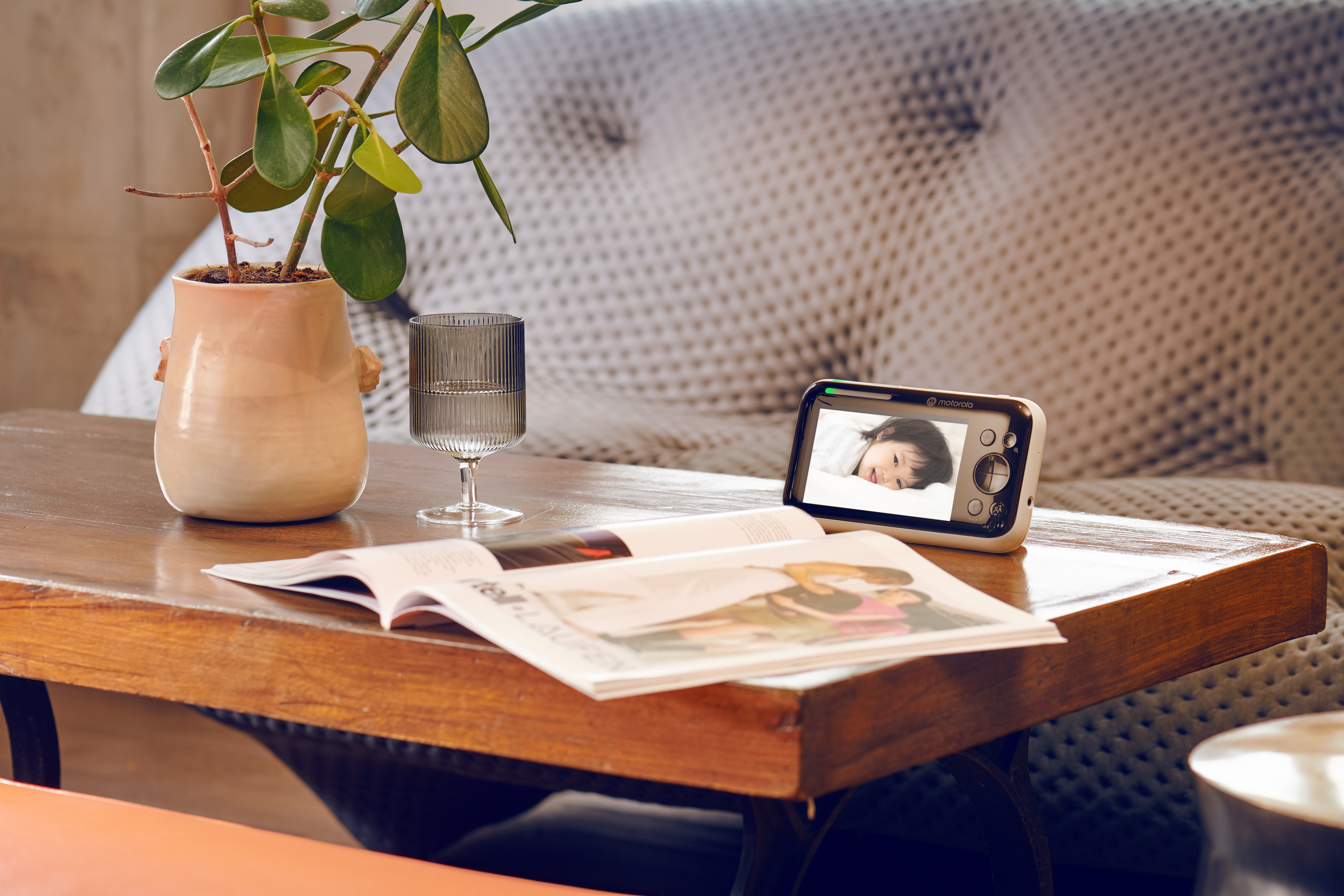 A baby monitor on a wooden table with a plant, a glass, and a magazine.