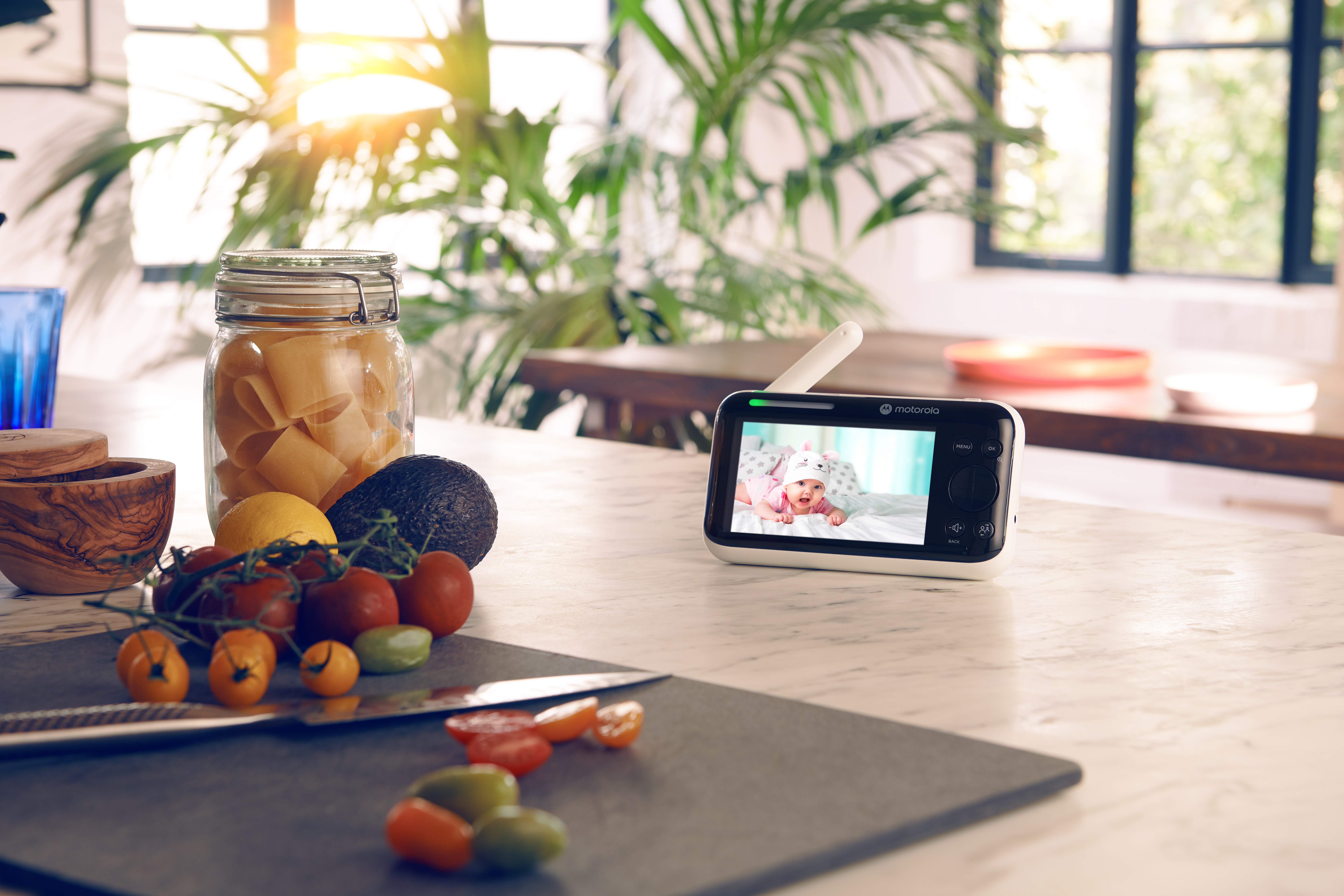Baby monitor che mostra un bambino. Il monitor è su un tavolo con cibo e una finestra sullo sfondo.