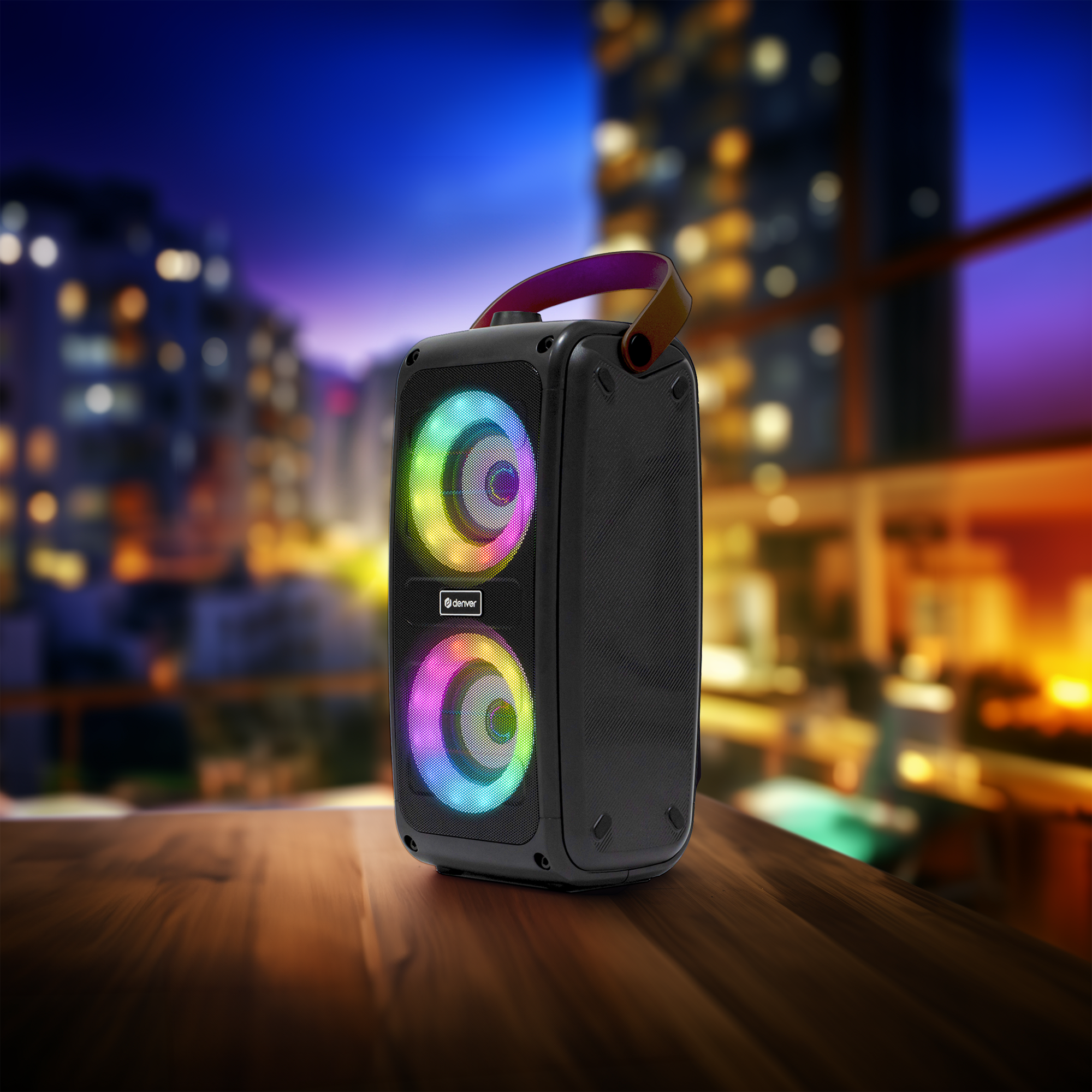 Un haut-parleur noir avec des lumières colorées est posé sur une surface en bois avec un paysage urbain en arrière-plan.