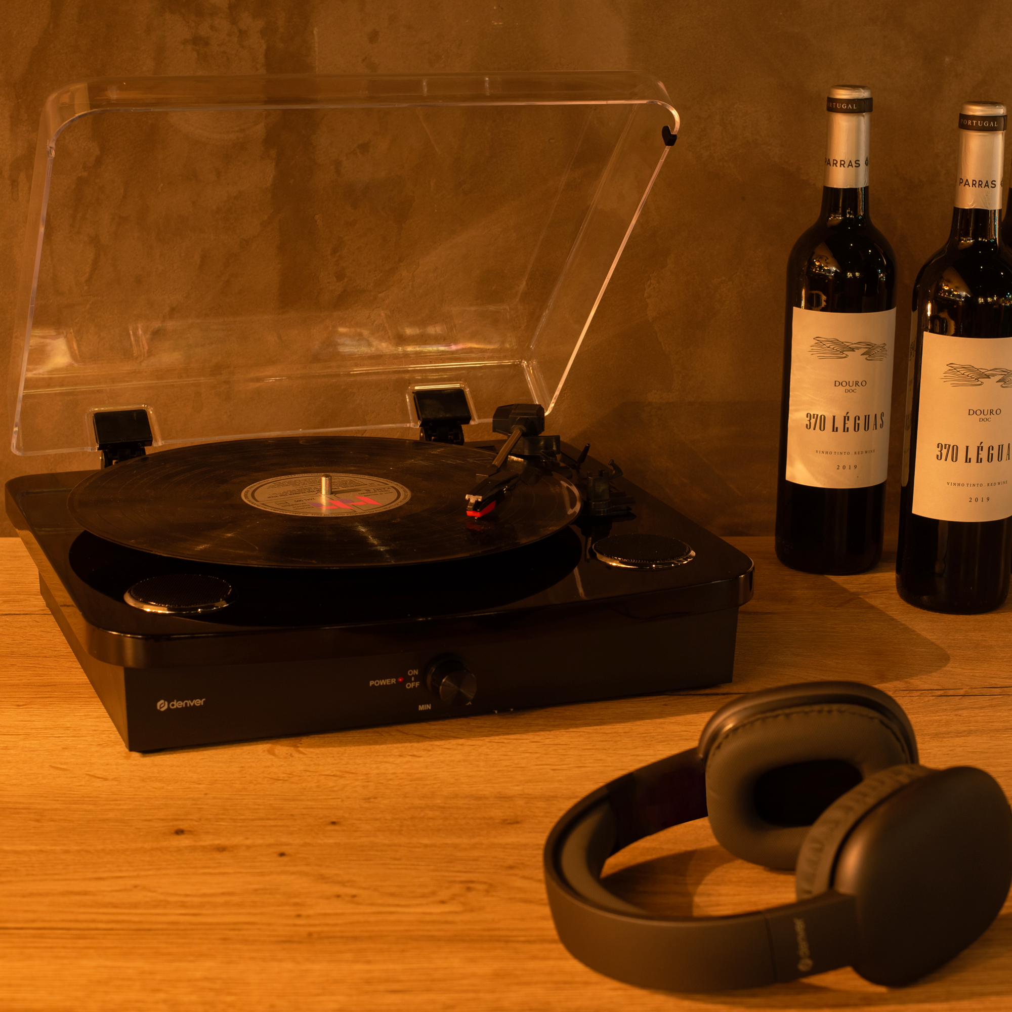 Gramofon z przezroczystą pokrywą. Dwie butelki czerwonego wina i słuchawki na drewnianej powierzchni.