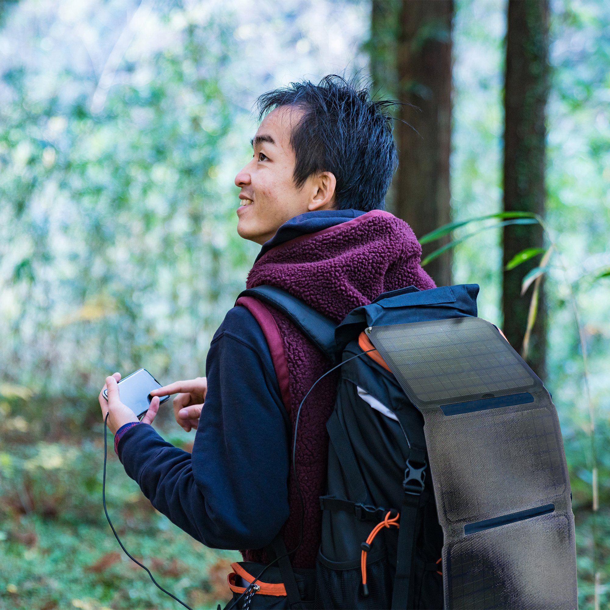 Mężczyzna w lesie z plecakiem. Używa telefonu podłączonego do panelu słonecznego.