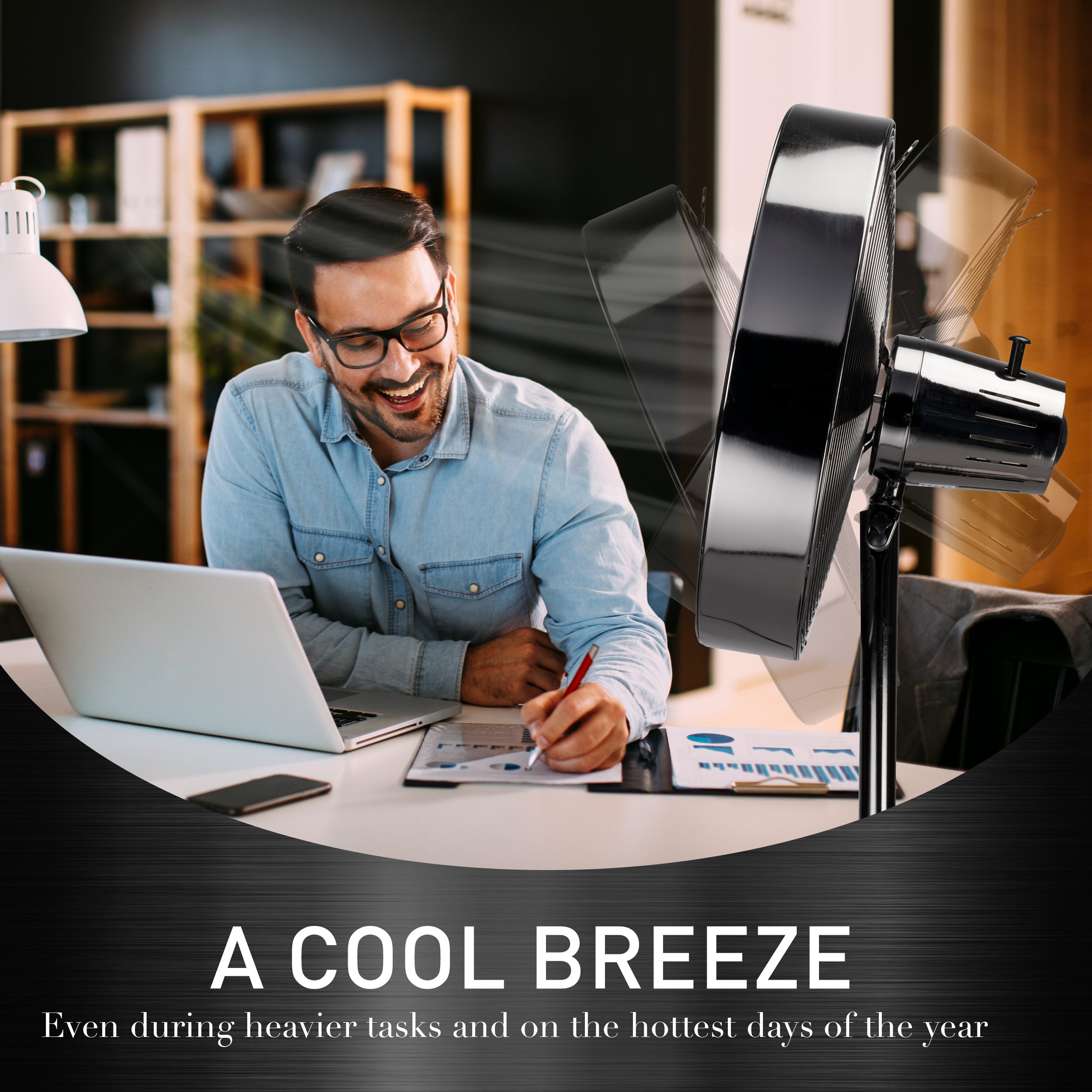 Man smiling while working at a desk with a laptop and a black oscillating fan.