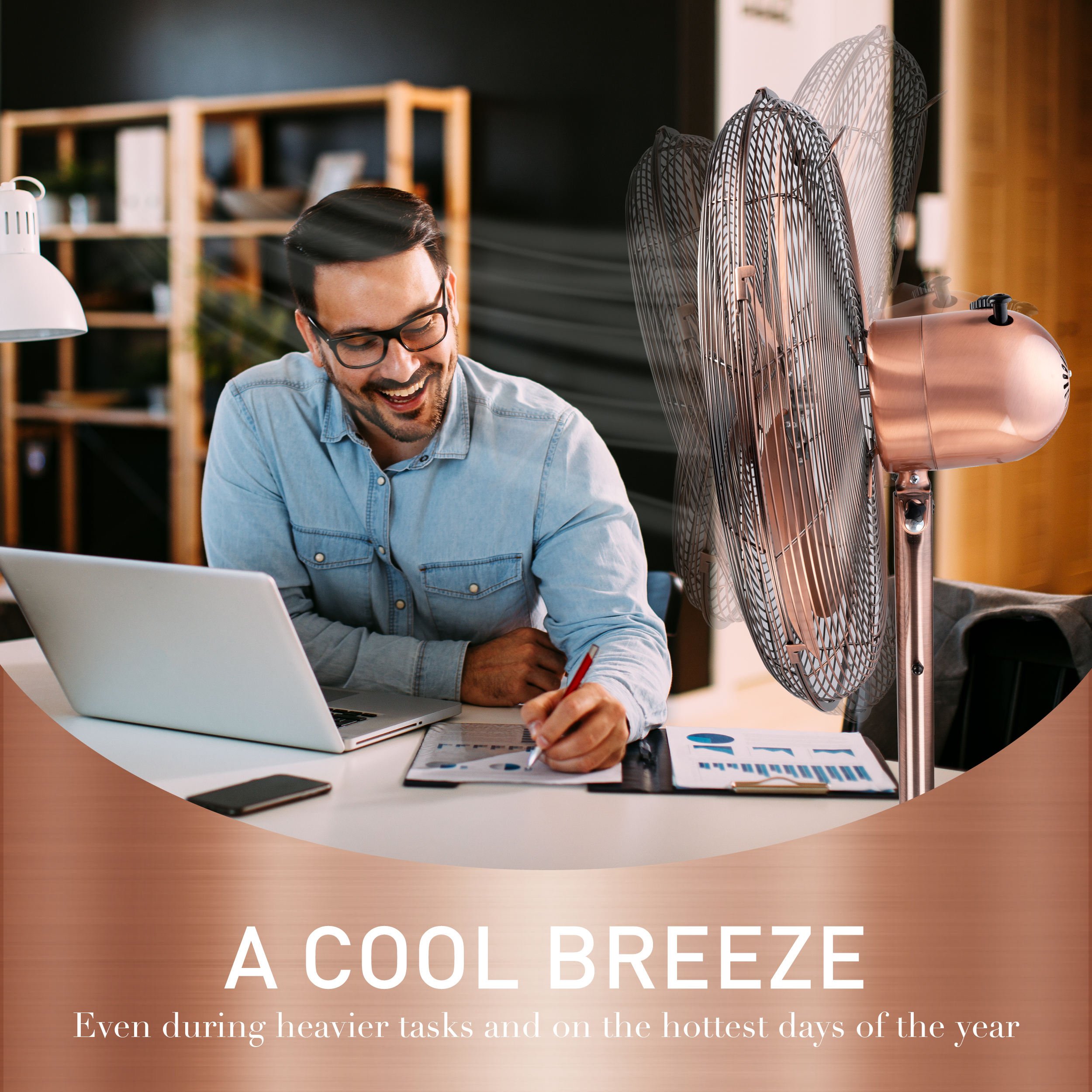Un homme avec des lunettes, souriant tout en travaillant sur un ordinateur portable. Un ventilateur en or rose souffle de l'air à proximité sur un bureau blanc.