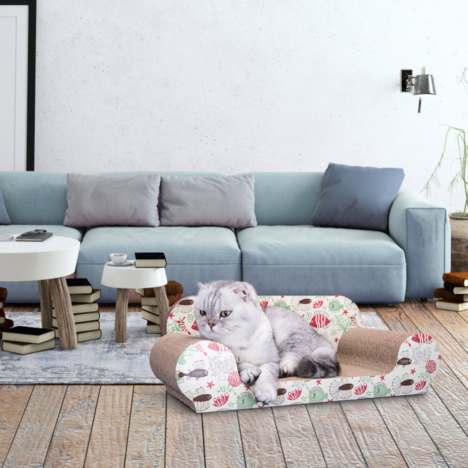 Un gato gris descansa en un sofá para gatos con un patrón de peces. Detrás, un sofá azul, una mesa blanca y libros.