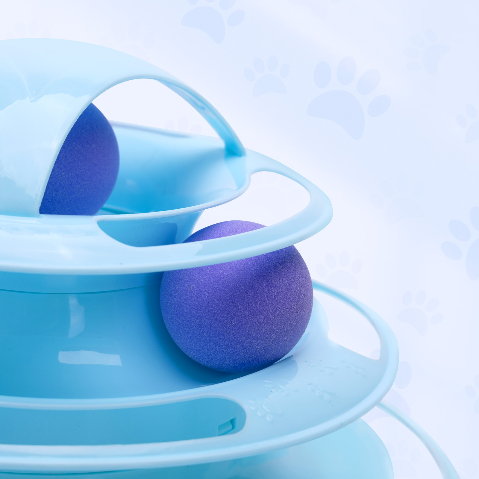 Juguete para gatos con dos bolas moradas sobre pistas azules. Fondo celeste con huellas de patas.