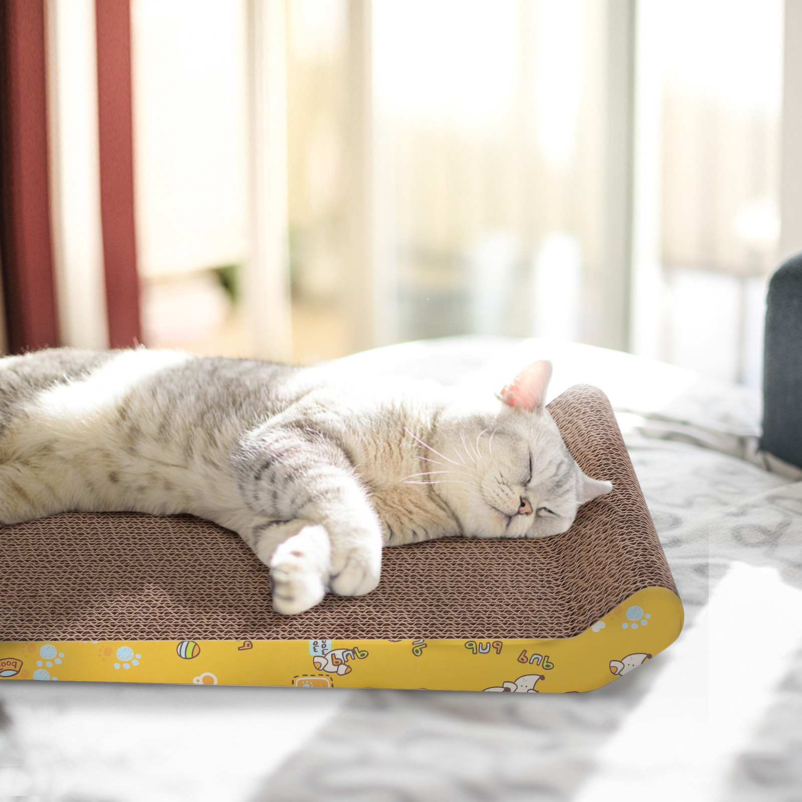 Un gato plateado durmiendo en un rascador amarillo, con una ventana de fondo.