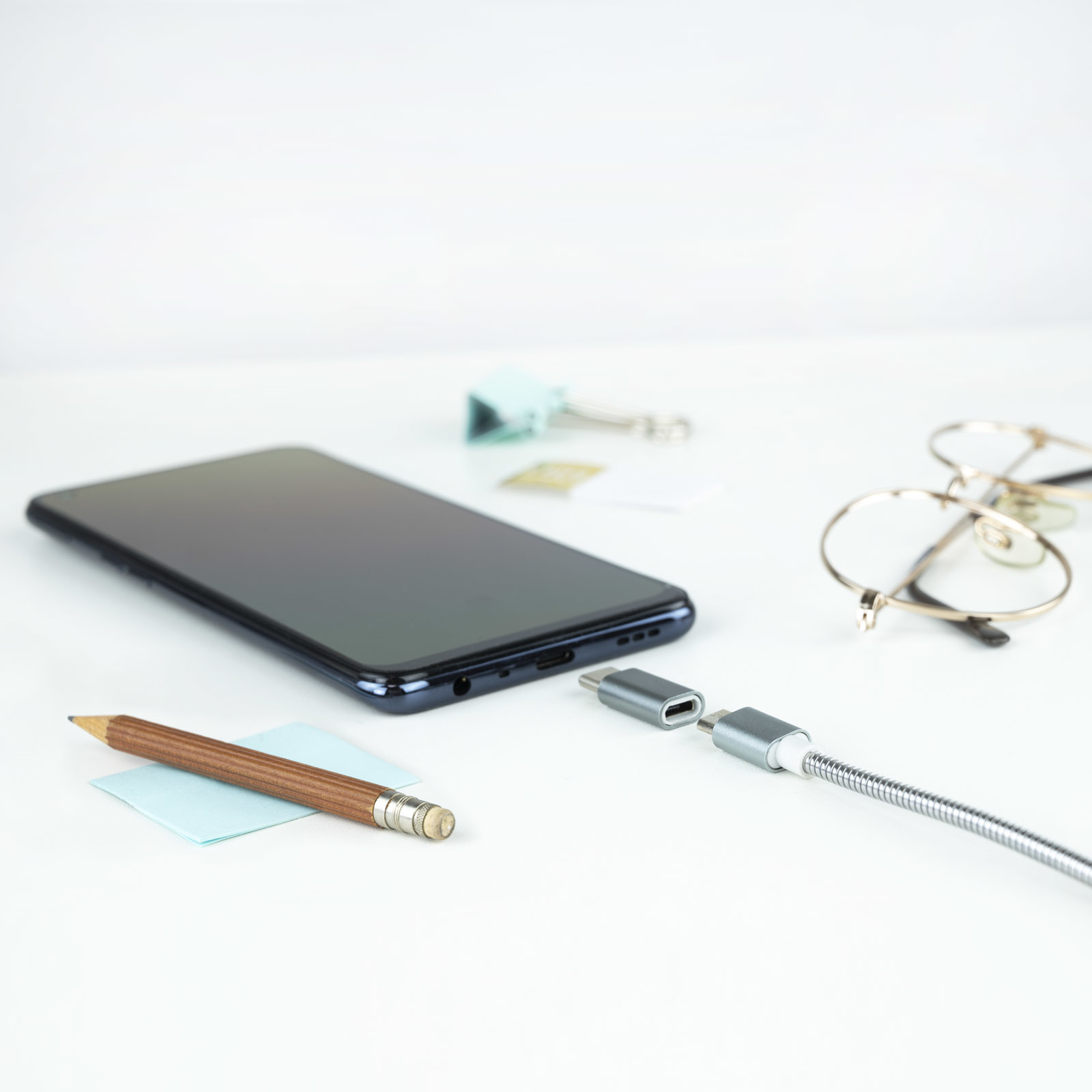 Smartphone, bolígrafo, nota adhesiva, gafas y cable de carga sobre una superficie blanca.