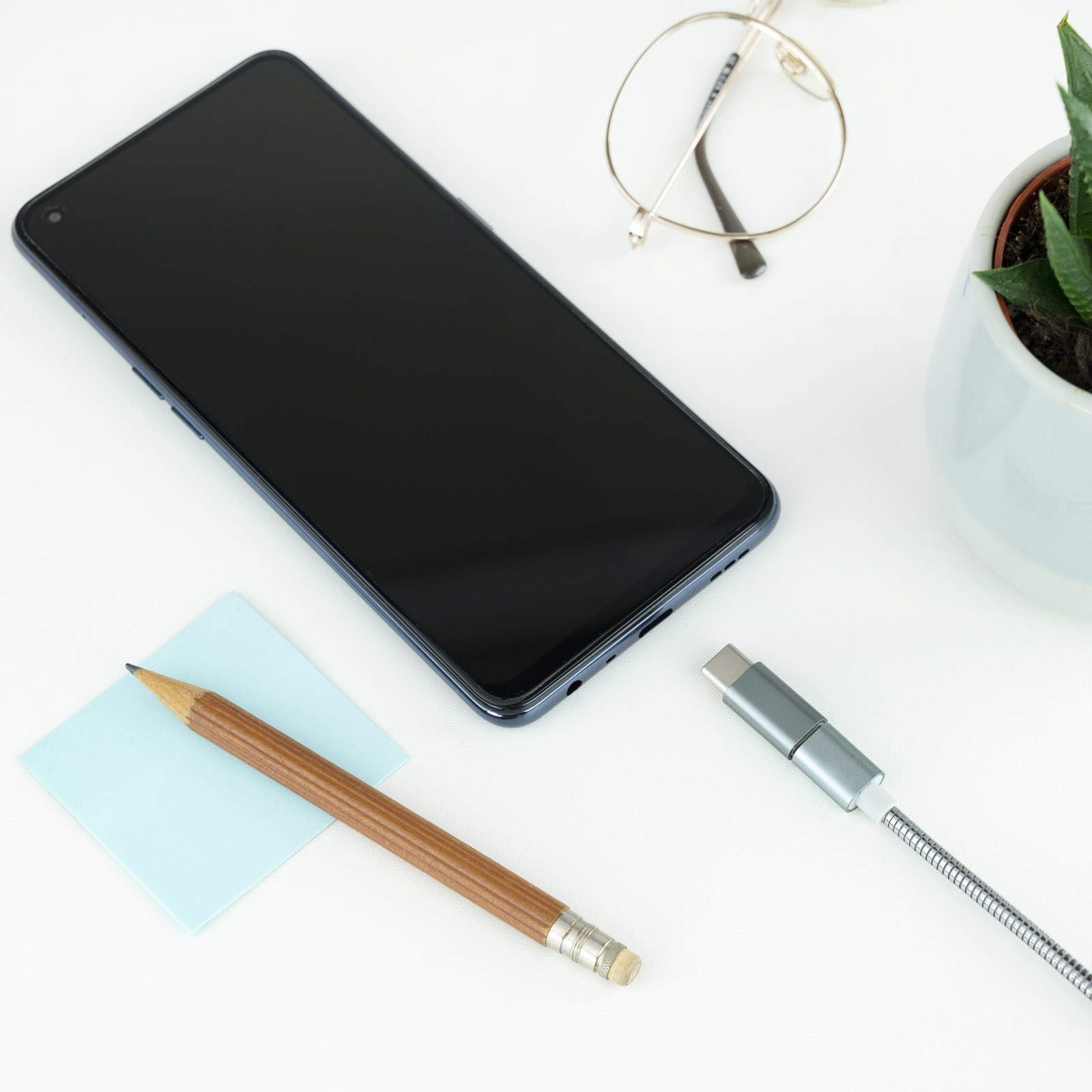 Un smartphone, cable, lápiz, gafas y una planta sobre una superficie blanca. Una configuración de oficina moderna.