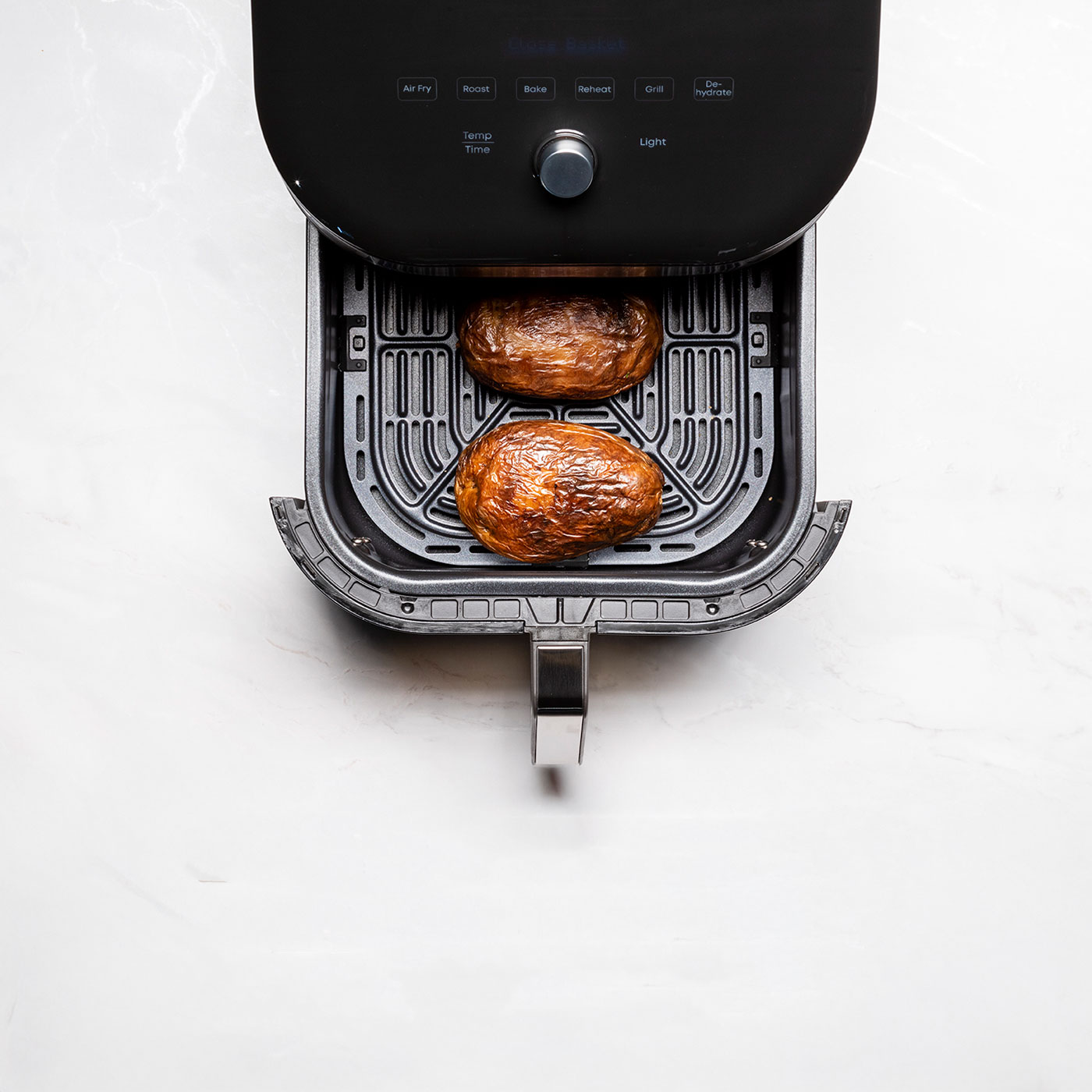 Widok z góry na frytkownicę powietrzną z dwoma ugotowanymi ziemniakami w środku, na białej powierzchni.