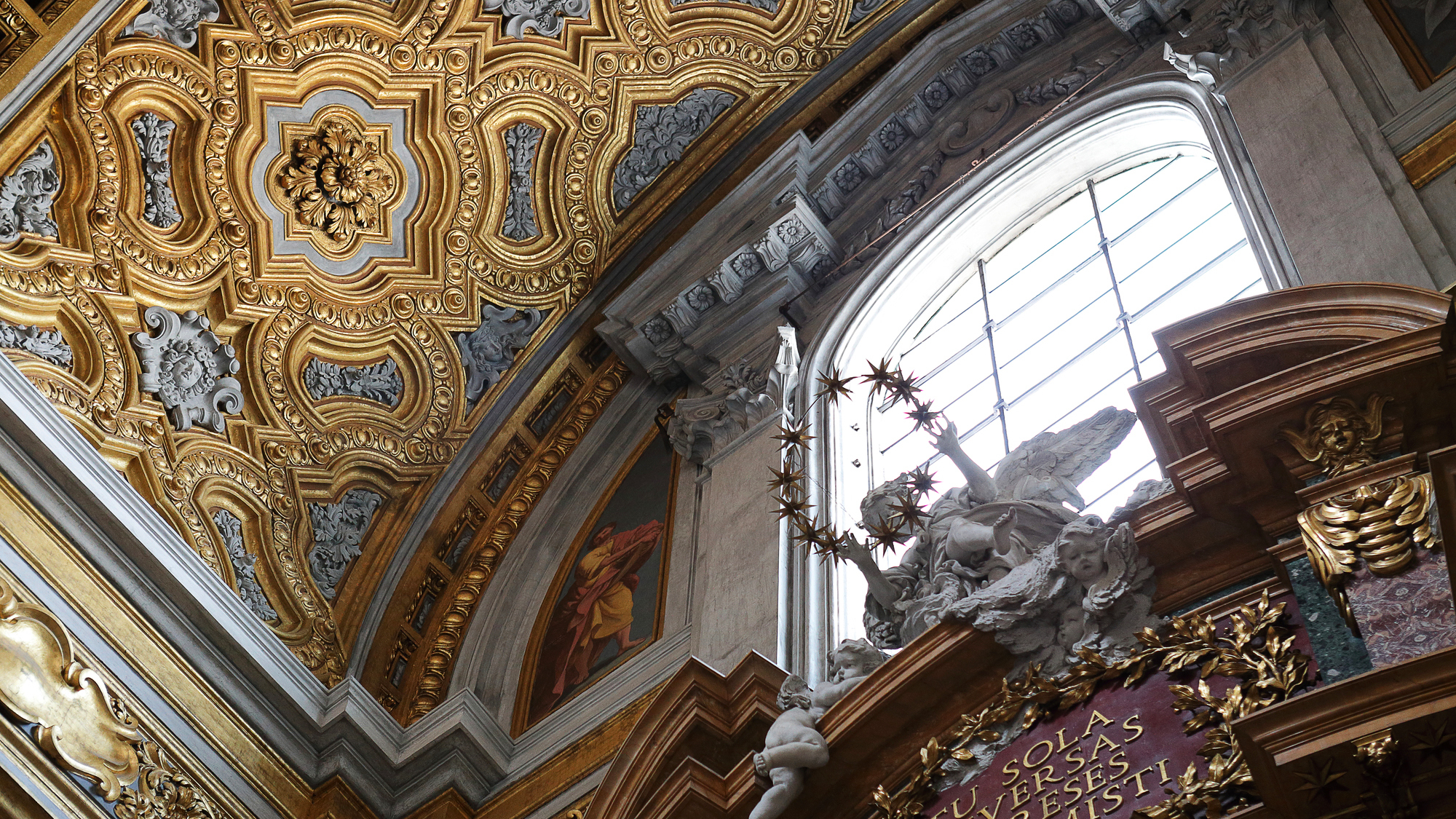 Soffitto dorato ornato con intagli dettagliati. Una finestra rivela una statua con angeli e un ornamento a forma di stella.