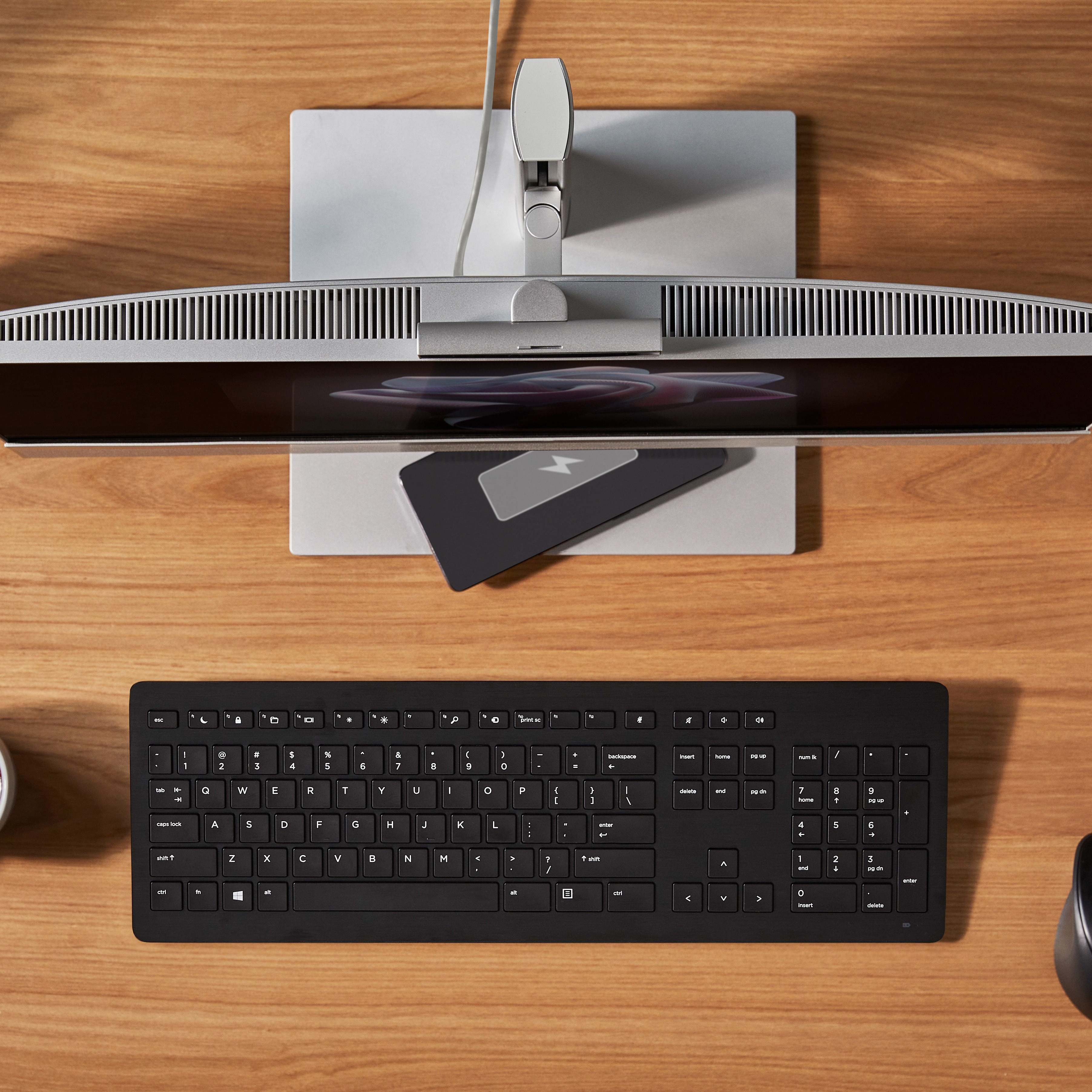 Widok z góry na biurko z monitorem, klawiaturą i małą podkładką ładującą.