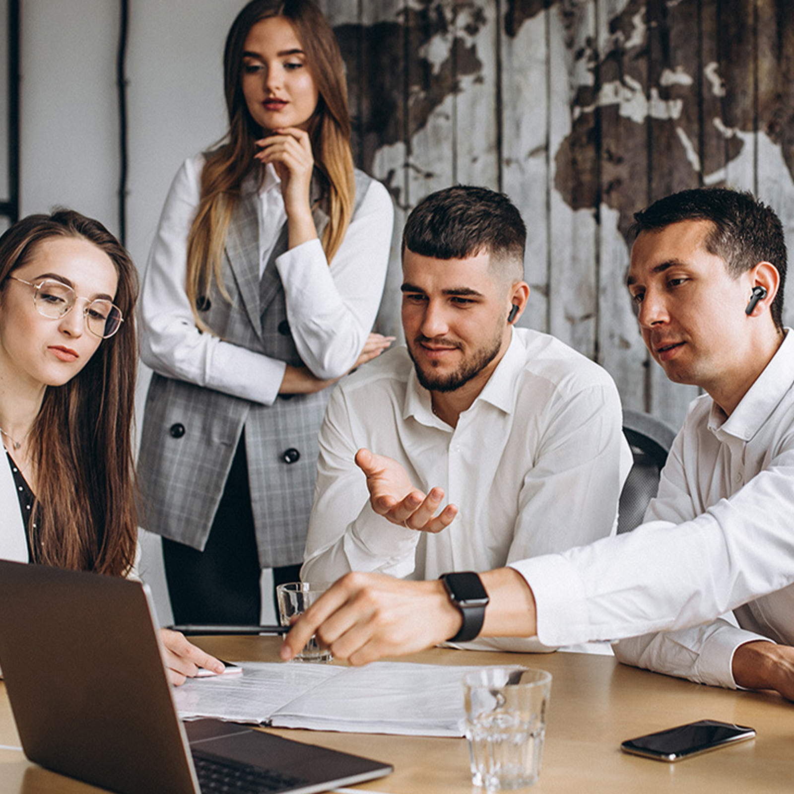 Vier Personen in einem Büro-Meeting. Einer zeigt auf ein Dokument. Im Hintergrund eine Weltkarte.