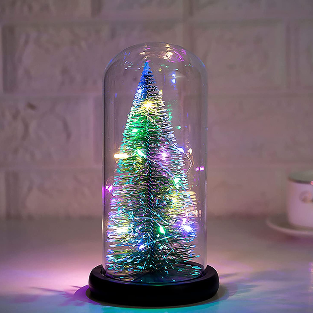 Weihnachtsbaum in einer Glaskuppel mit bunten Lichtern, auf weißer Oberfläche.