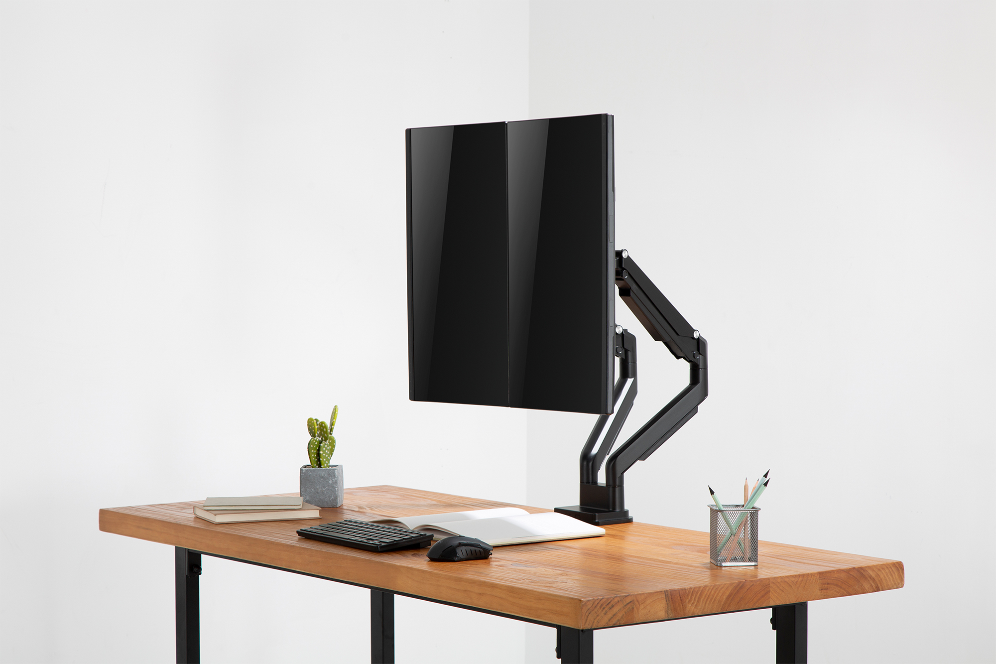 Dwa czarne monitory zamontowane na drewnianym biurku. Widoczny jest także kaktus, klawiatura, mysz i długopisy.