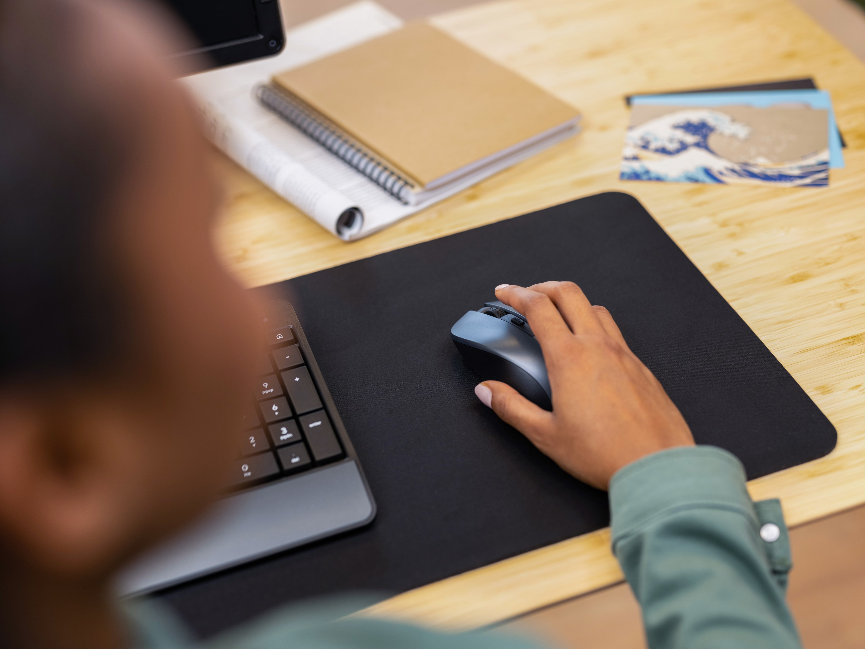 Ręka osoby używającej myszy na czarnej podkładce. Obok notatnik i karty.