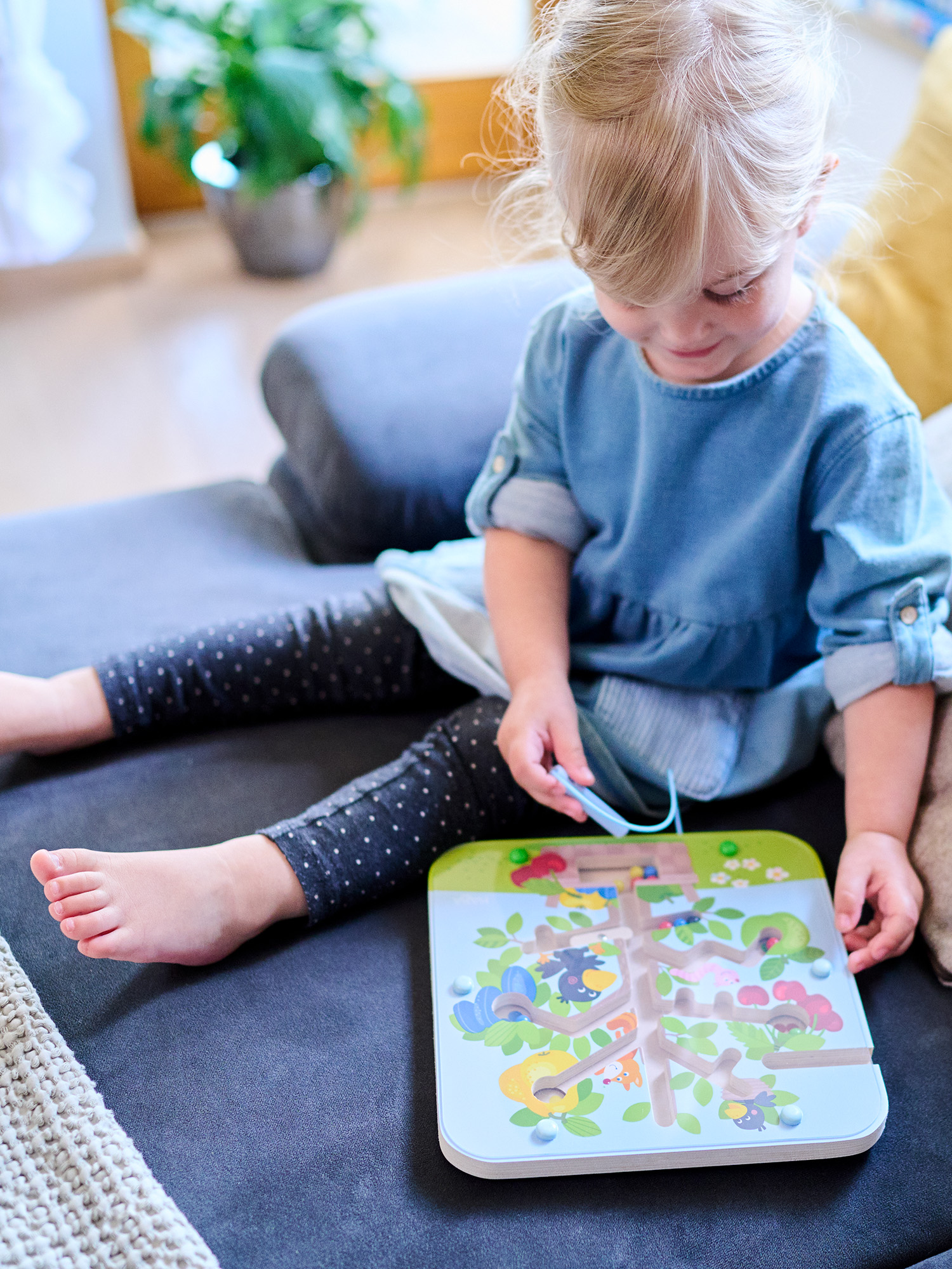 Ein kleines Mädchen spielt mit einem Holzbrettspiel, während es auf einem grauen Sofa sitzt.