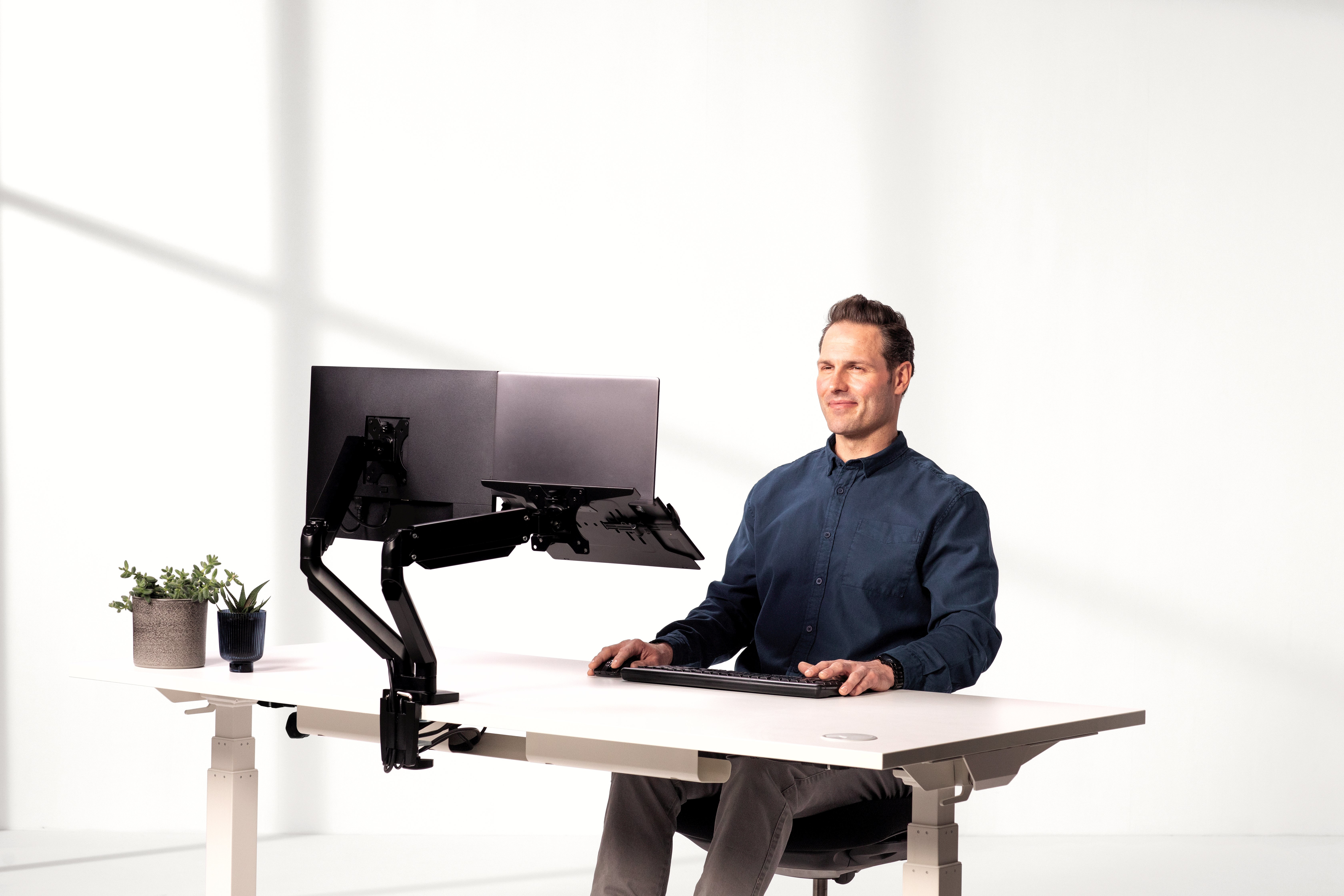 Mężczyzna siedzi przy białym biurku z dwoma monitorami, klawiaturą i myszką. Otoczenie biurowe z roślinami.