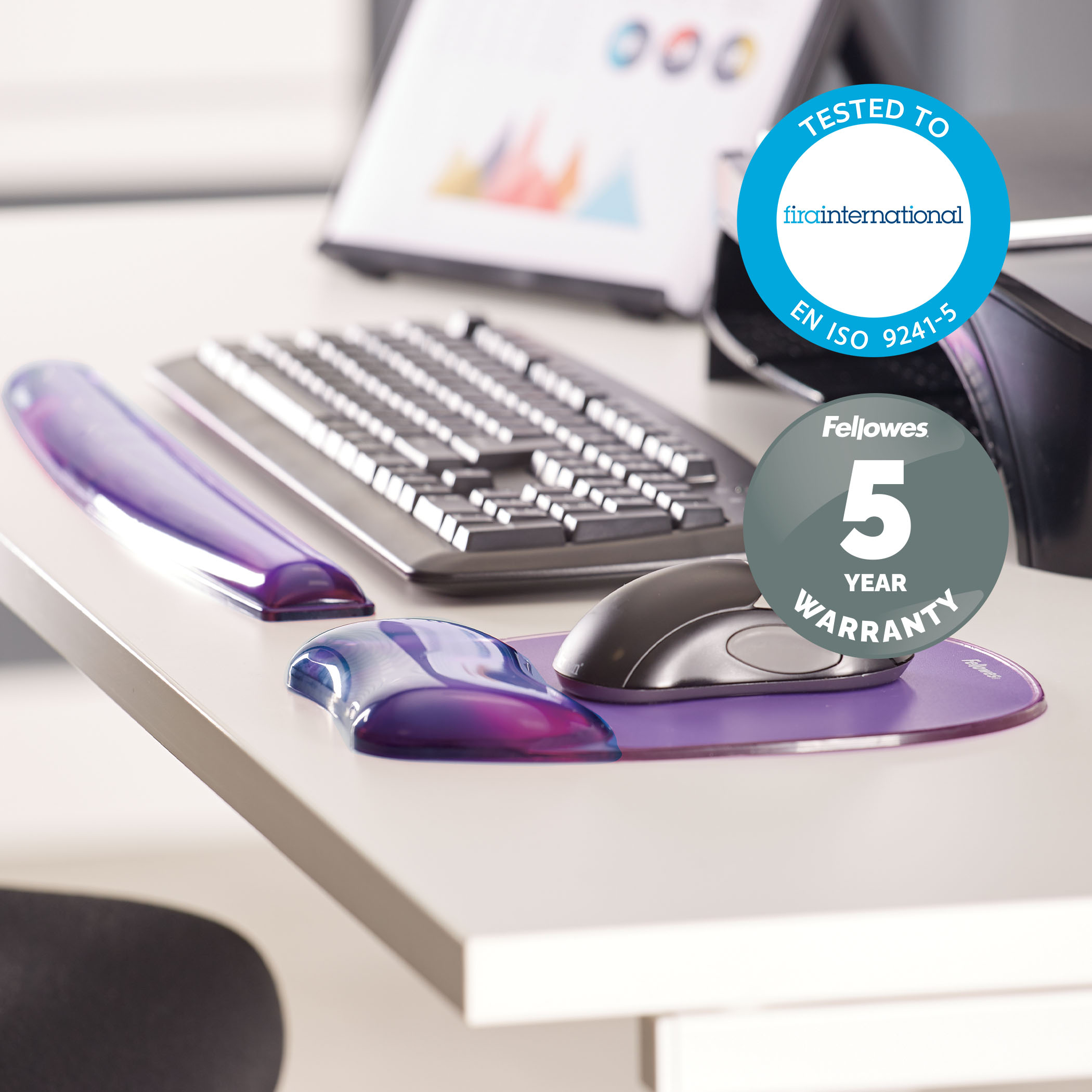 Klawiatura, mysz i podpórka pod nadgarstek na białym biurku. Dwa loga: 'Testowane zgodnie z EN ISO 9241-5'.