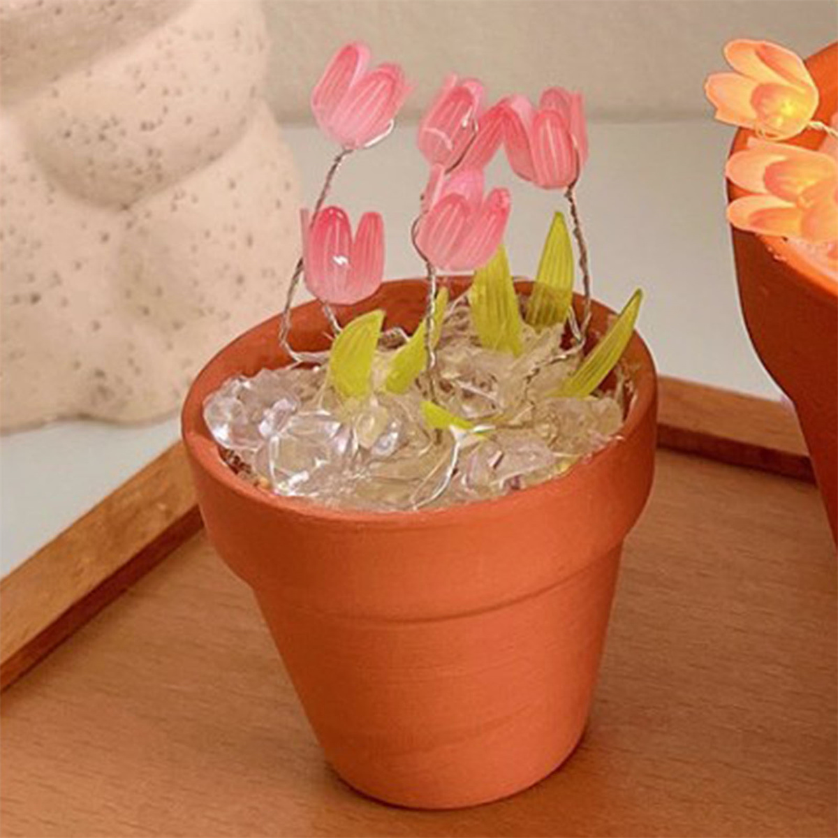 Ein kleiner Terrakottatopf mit rosa tulpenartigen Blüten und klaren Steinen auf einem Holztablett.