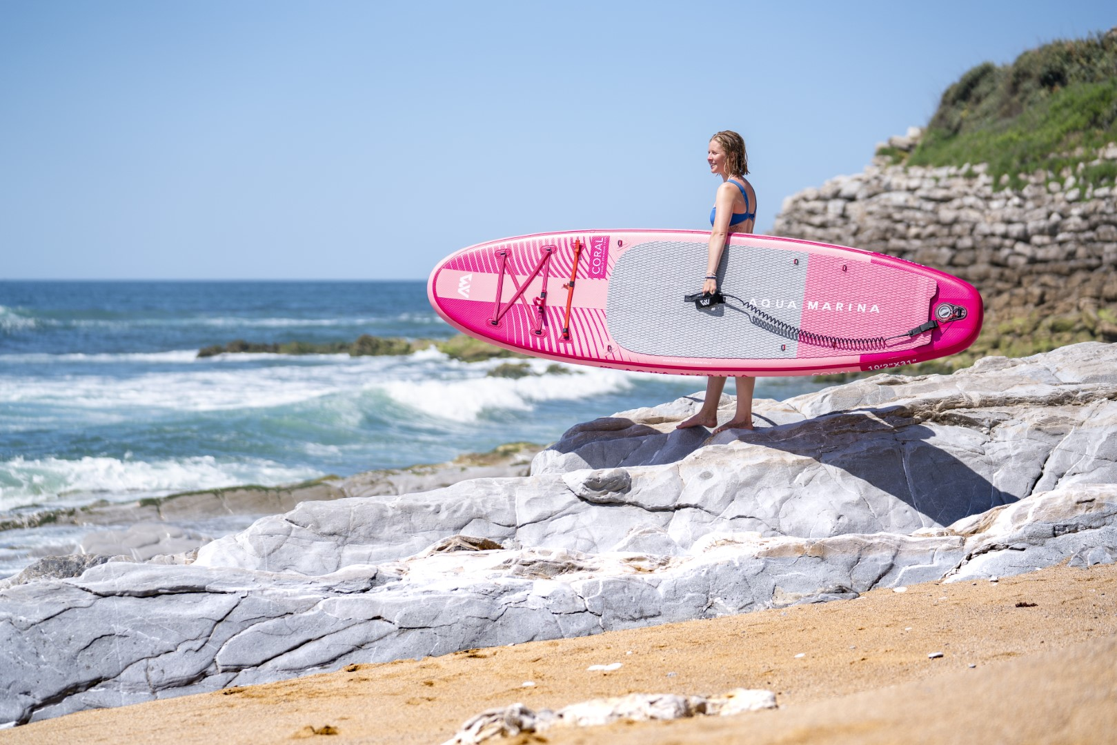 Eine Frau mit einem pinken Surfbrett an einem Strand, mit Felsen, Sand und Wellen im Hintergrund.