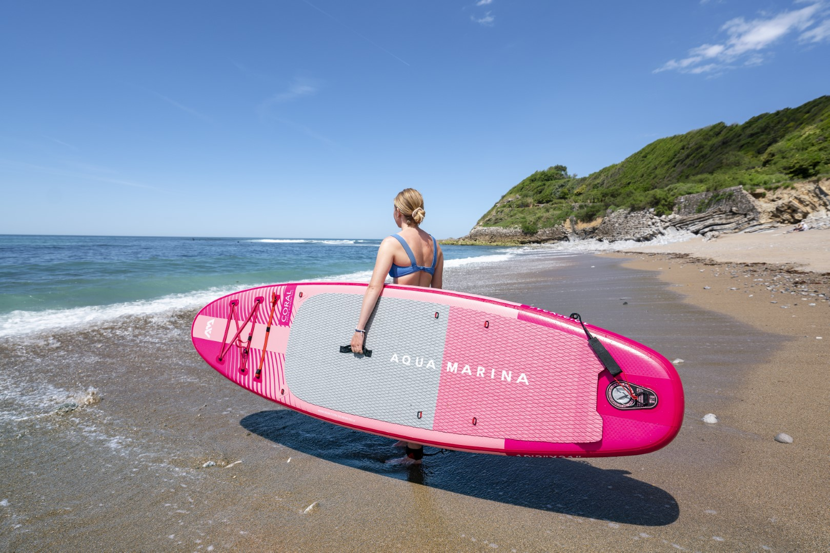 Eine Frau, die ein pinkes Paddleboard an einem Sandstrand mit blauem Meer und Himmel hält.