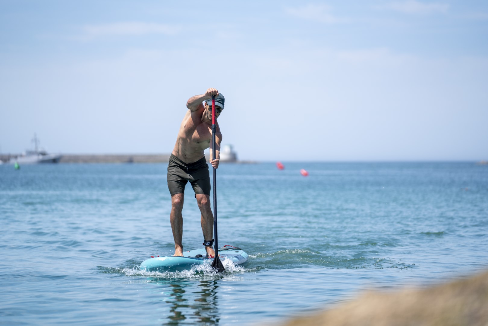 Mężczyzna uprawiający paddleboarding na niebieskiej desce, w niebieskiej wodzie, w słoneczny dzień.