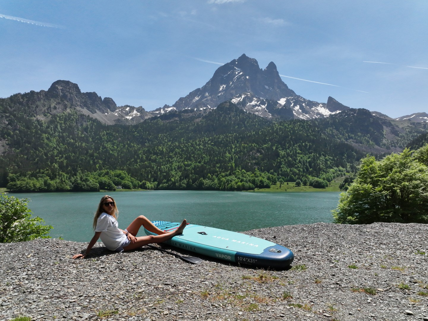 Kobieta siedzi na desce do wiosłowania na jeziorze. W tle góra. Niebo jest niebieskie.