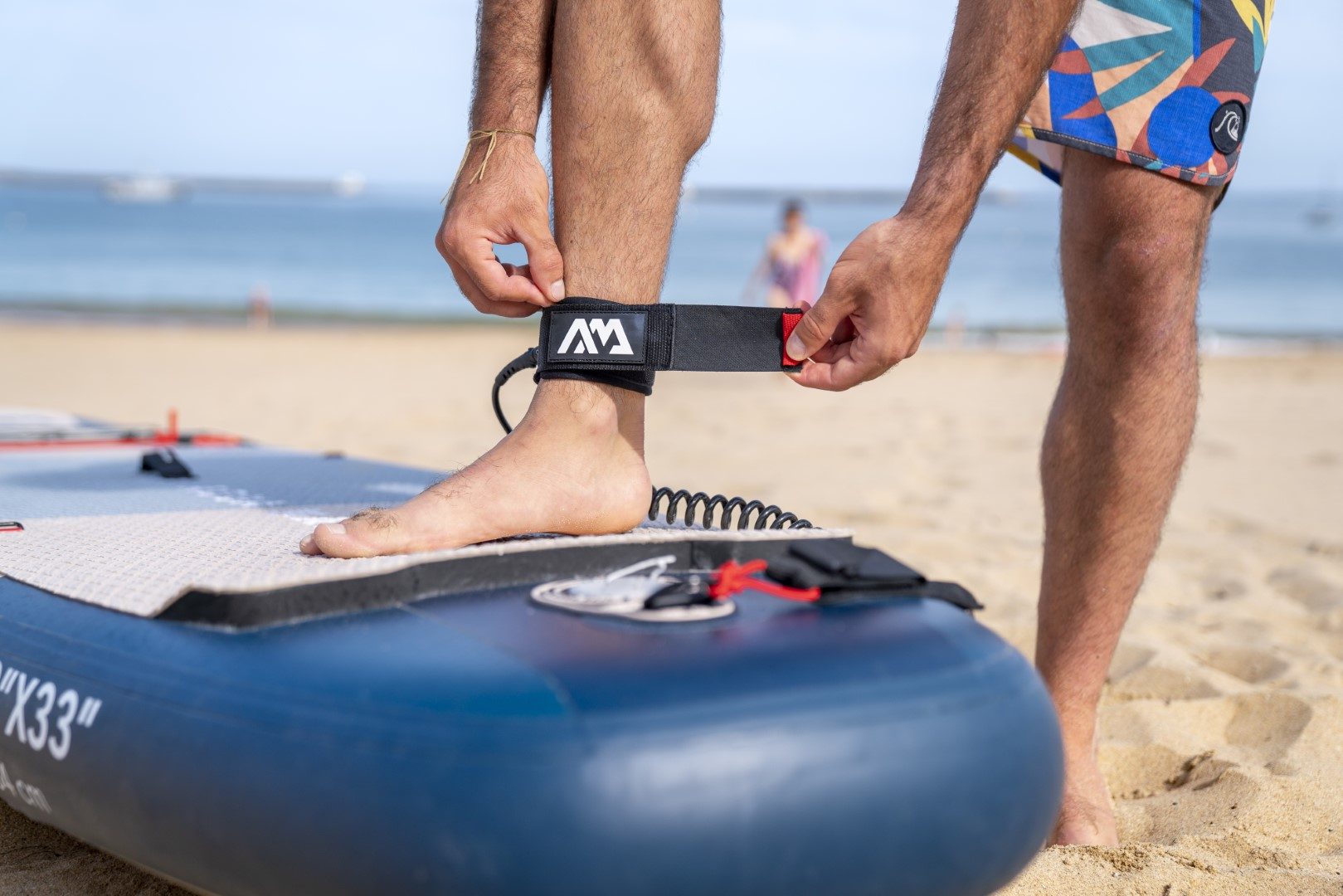 Osoba mocująca czarną smycz do kostki do niebieskiej deski SUP na piaszczystej plaży. Słoneczny dzień.