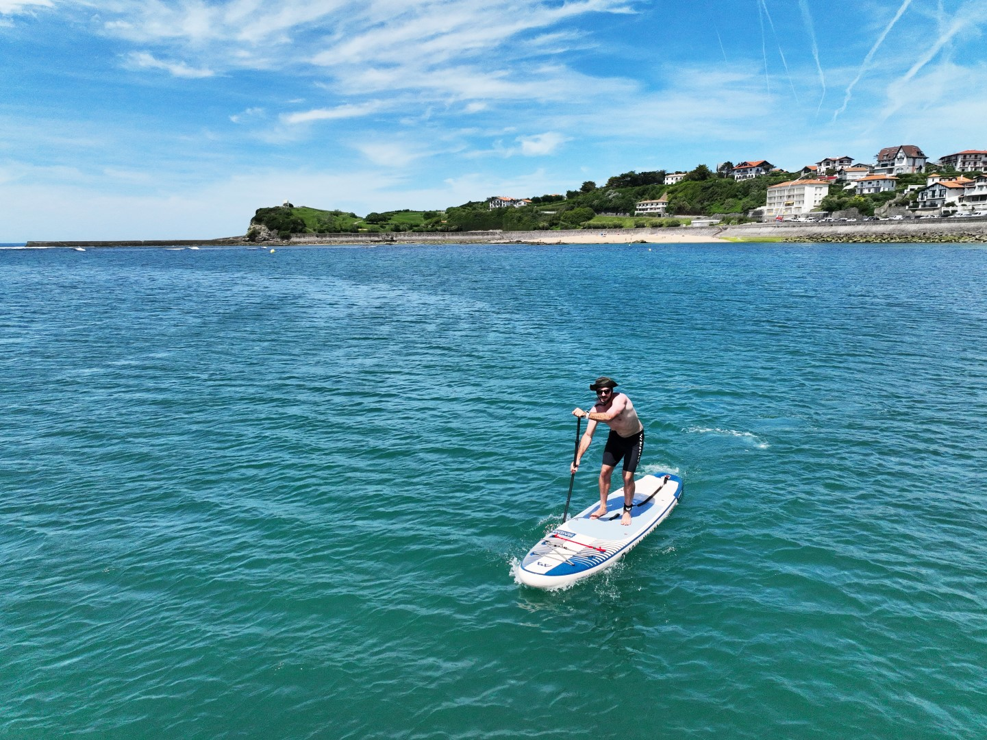 Mężczyzna na paddleboardzie na niebiesko-zielonej wodzie, domy i błękitne niebo w tle.