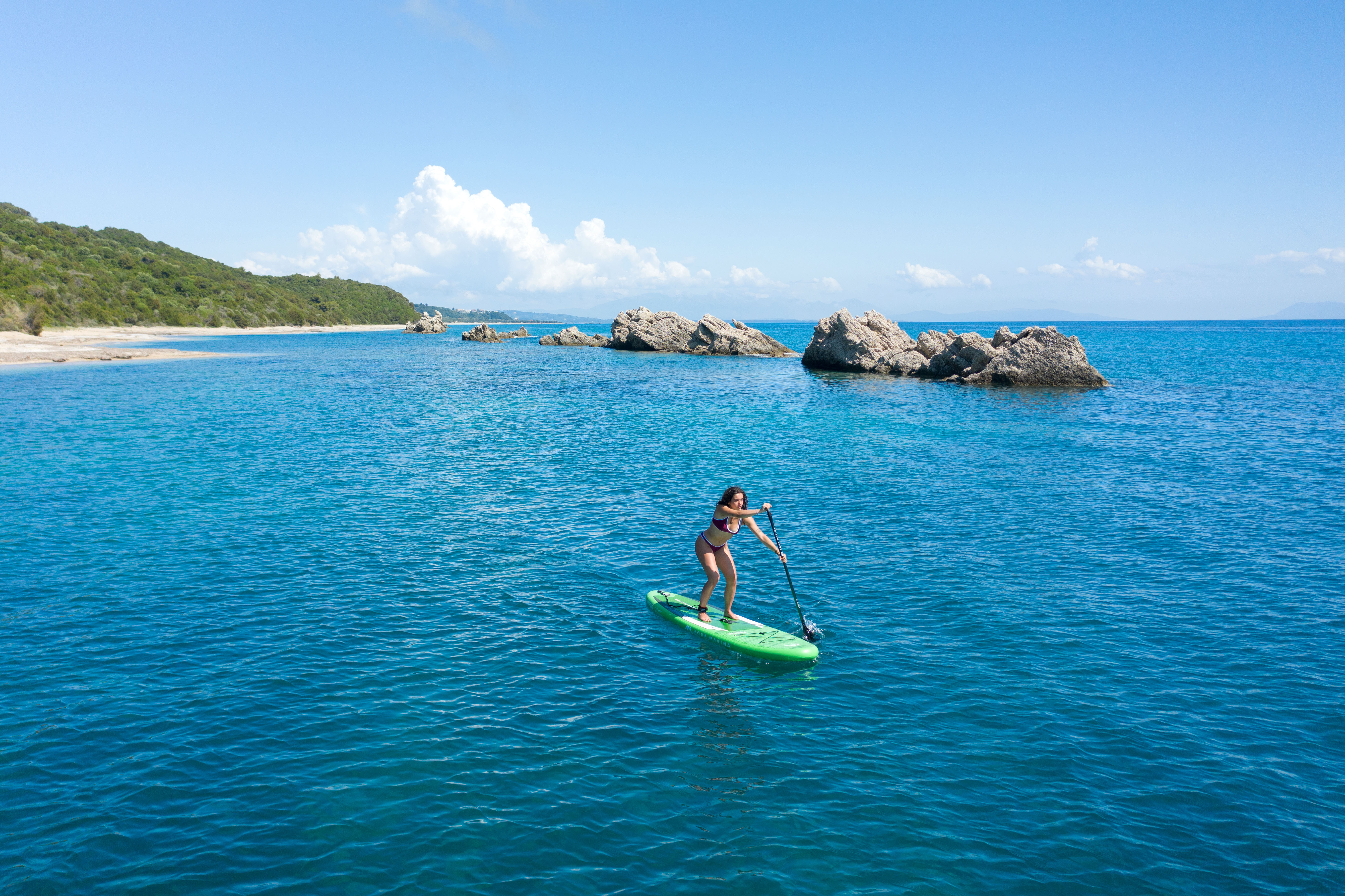 Frau beim Stand-Up-Paddling auf blauem Wasser, mit grünem Board und Felsen in der Ferne.