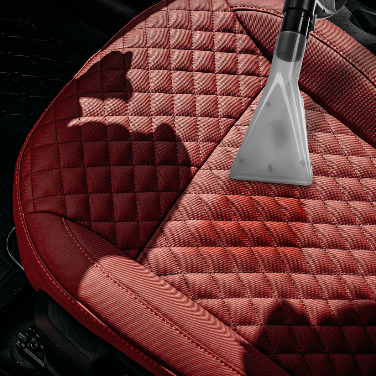 Zbliżenie czerwonego fotela samochodowego czyszczonego dyszą odkurzacza. Siedzisko ma diamentowe przeszycia.