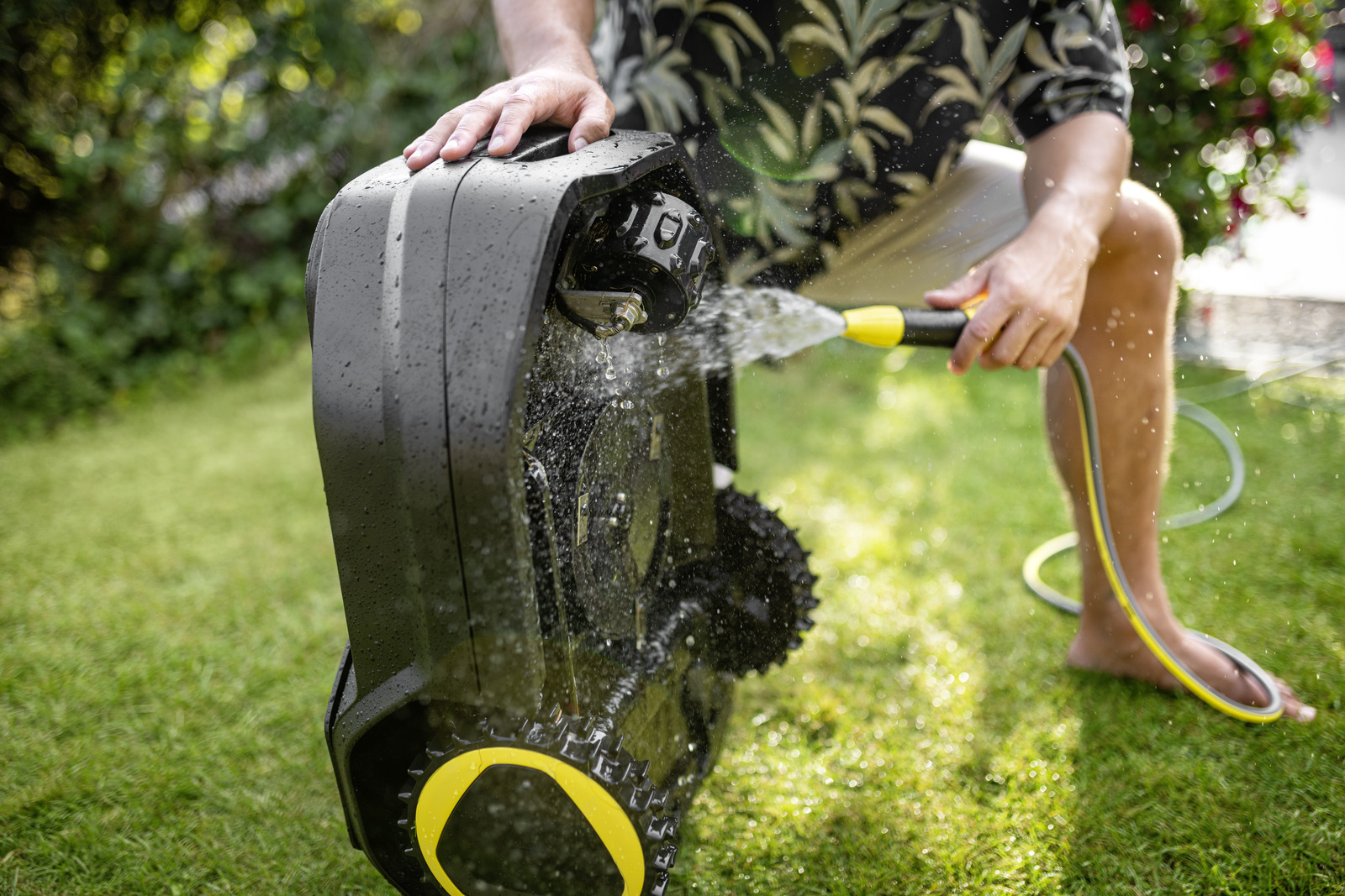 Osoba myjąca czarną kosiarkę automatyczną wężem na zielonym trawniku.