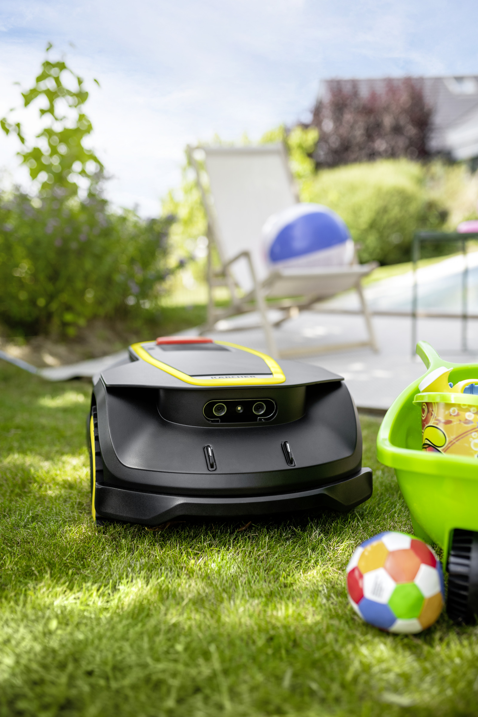 Robot koszący na trawie. Na pierwszym planie: piłki i zielony wózek. Tło: dom, leżak.