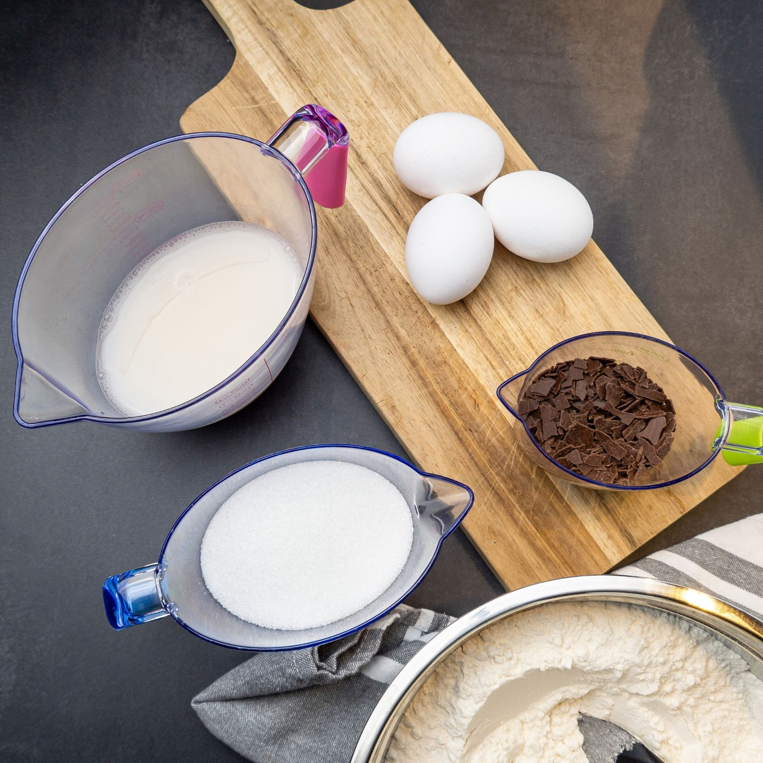 Zutaten zum Backen: Milch in einem Krug, Schokostückchen, Zucker und Eier auf einem Holzbrett.