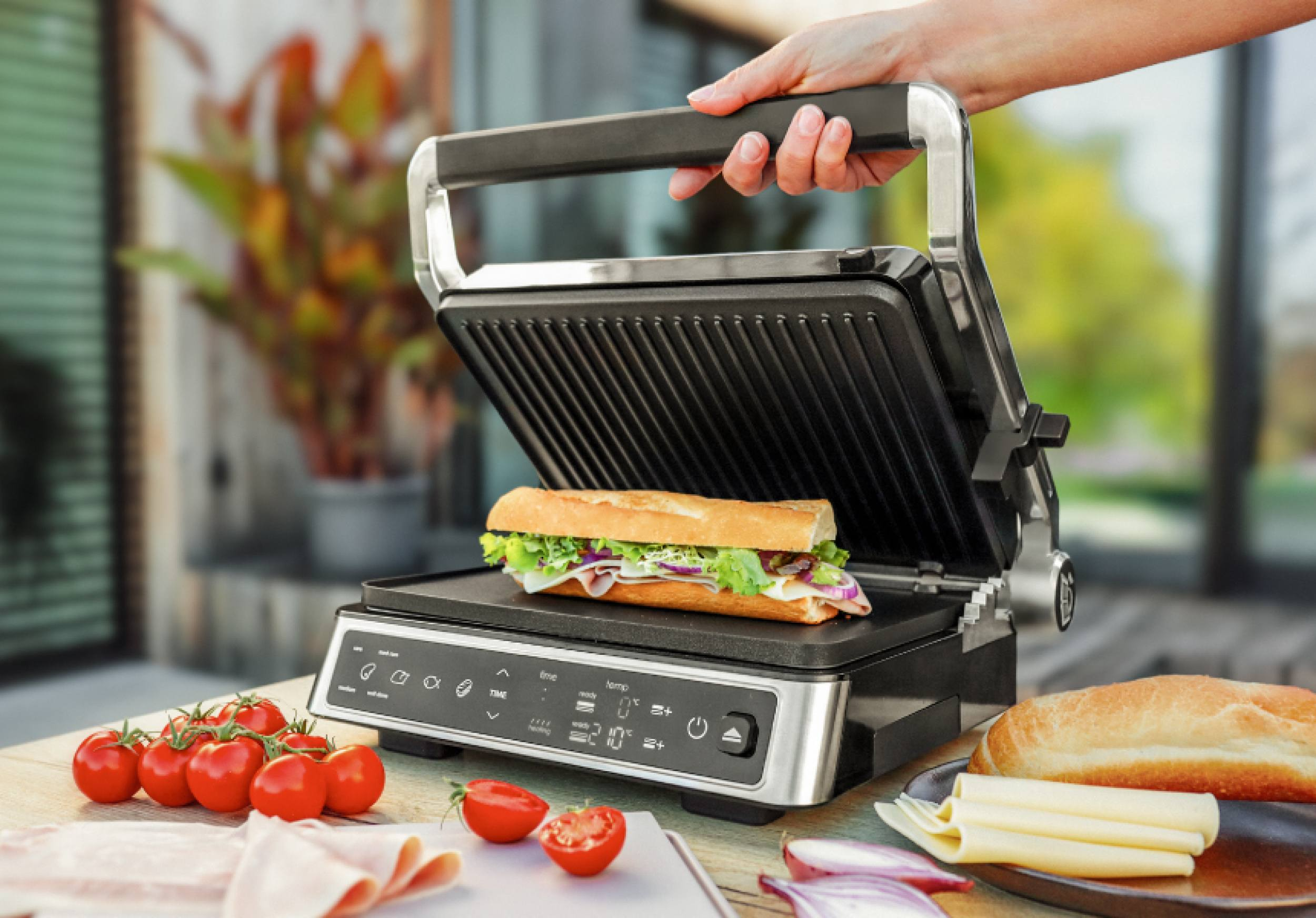 Sandwich auf einem Grill, Tomaten und Brot. Die Hand einer Frau hält den Grill. Holztisch.