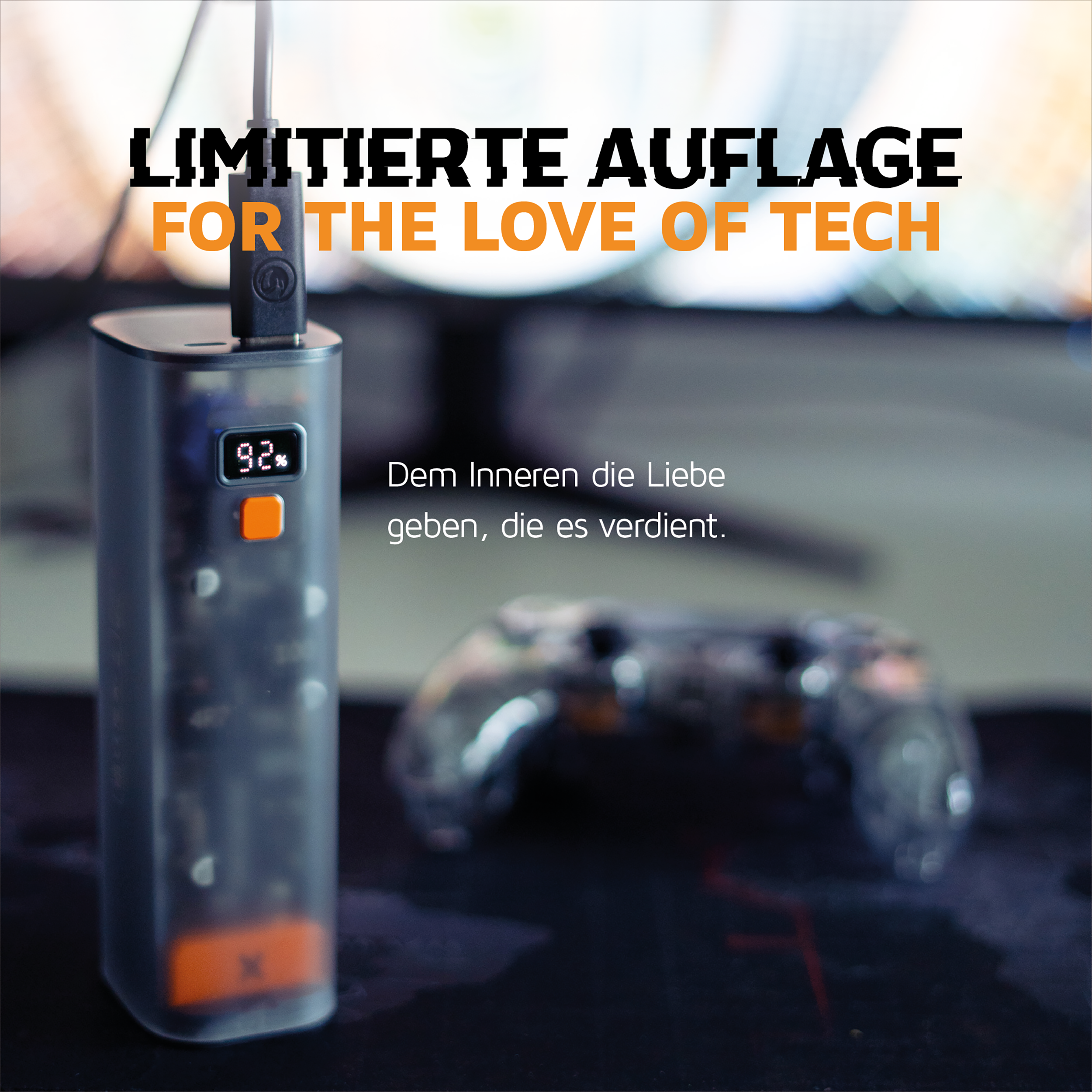 Eine transparente Powerbank mit orangefarbenem Knopf, die ein schwarzes Kabel auflädt. Ein Controller liegt im Hintergrund.