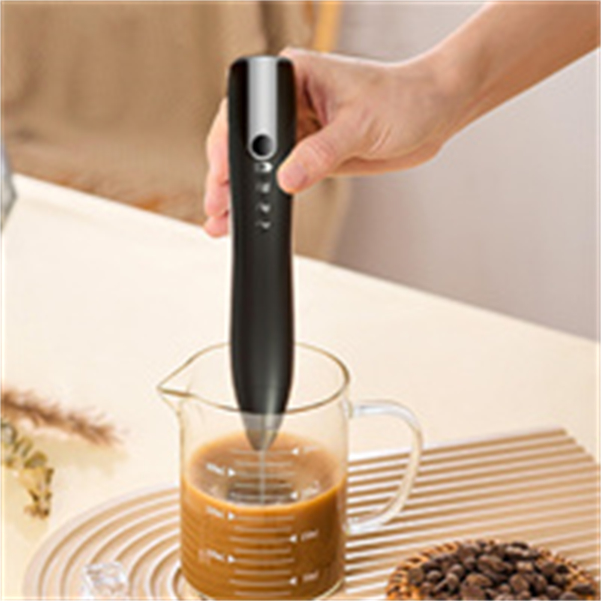 Hand hält einen schwarzen Milchaufschäumer über einem Glas mit Kaffee. Kaffeebohnen an der Seite.