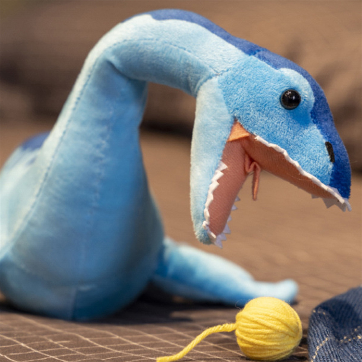 Blaues Plüschtier-Dinosaurier mit offenem Mund, mit Wollknäuel davor. Braune Oberfläche im Hintergrund.