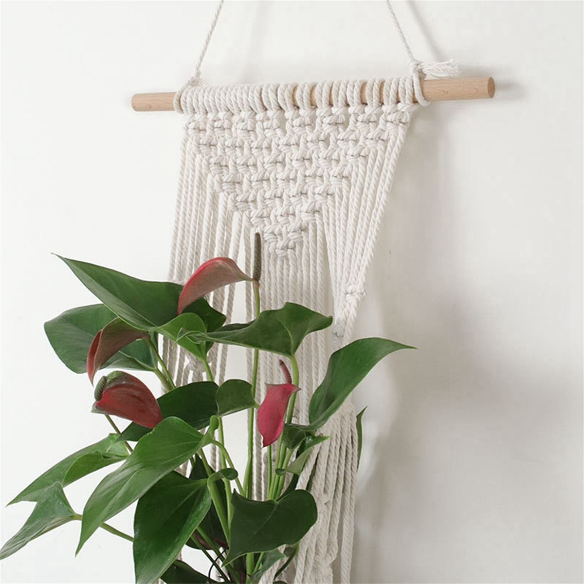 Makramee-Pflanzenaufhänger mit grünen Blättern und roten Blüten, an einer weißen Wand hängend.